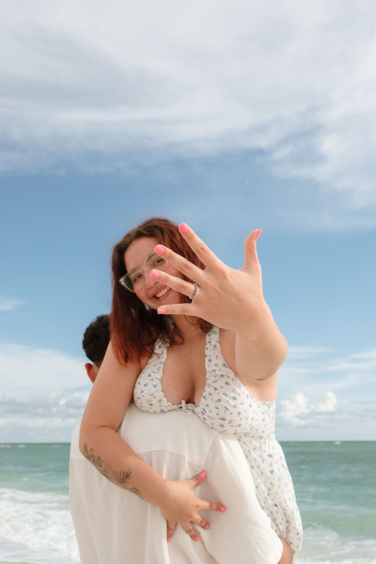 Woman showing engagement ring on beach while partner embraces her, ocean and blue sky in background.