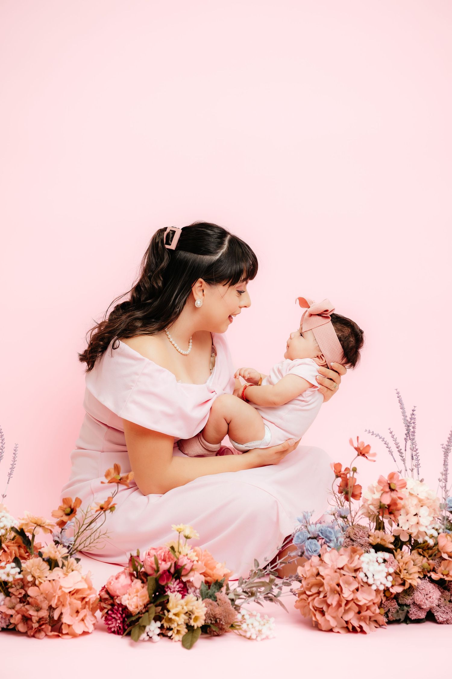 A mother in a white dress holds her newborn baby surrounded by colorful flowers on a pink background.