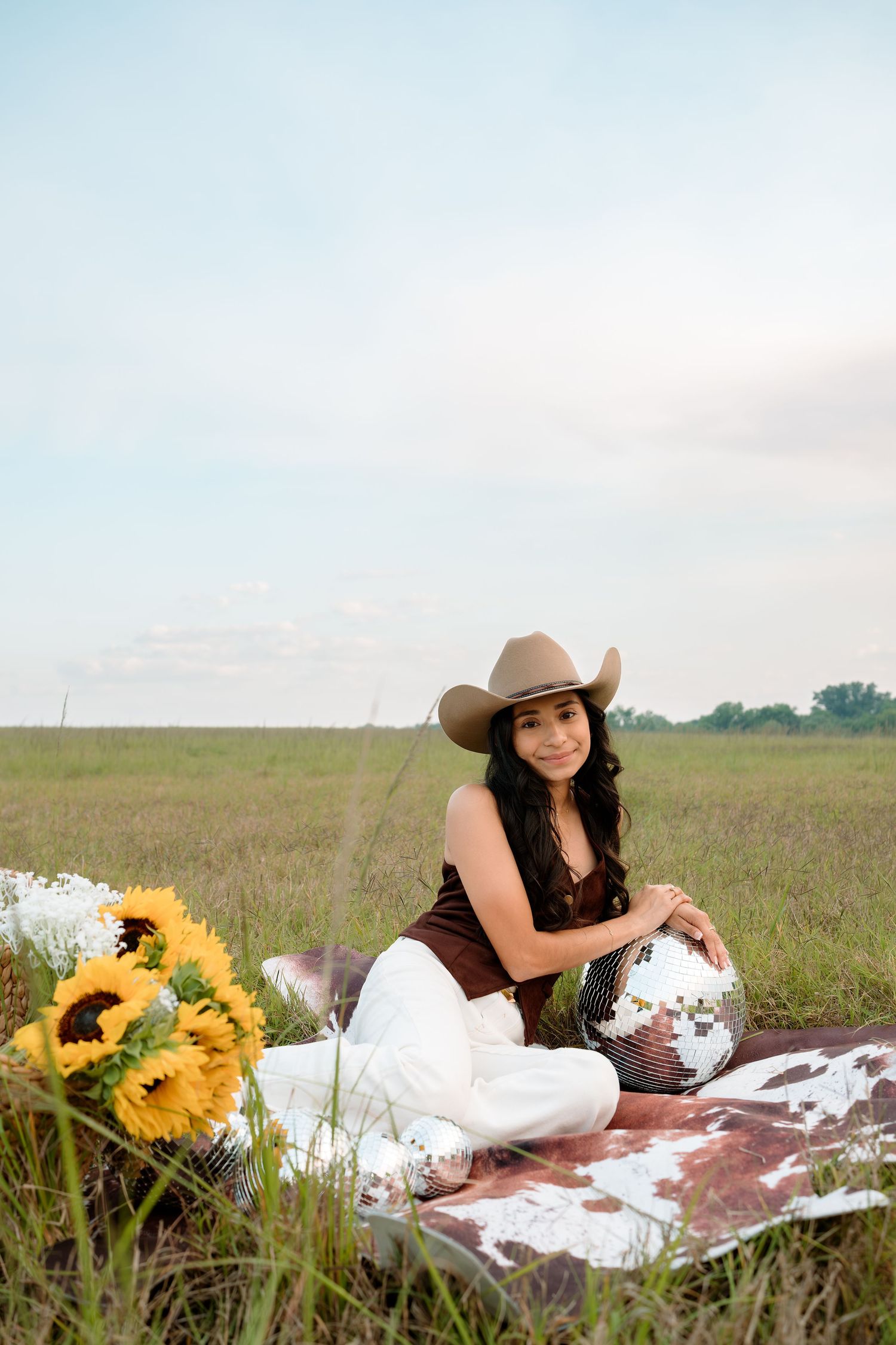 Young woman in cowboy hat sits cross-legged in a field with sunflowers and a disco ball on a cowhide blanket.
