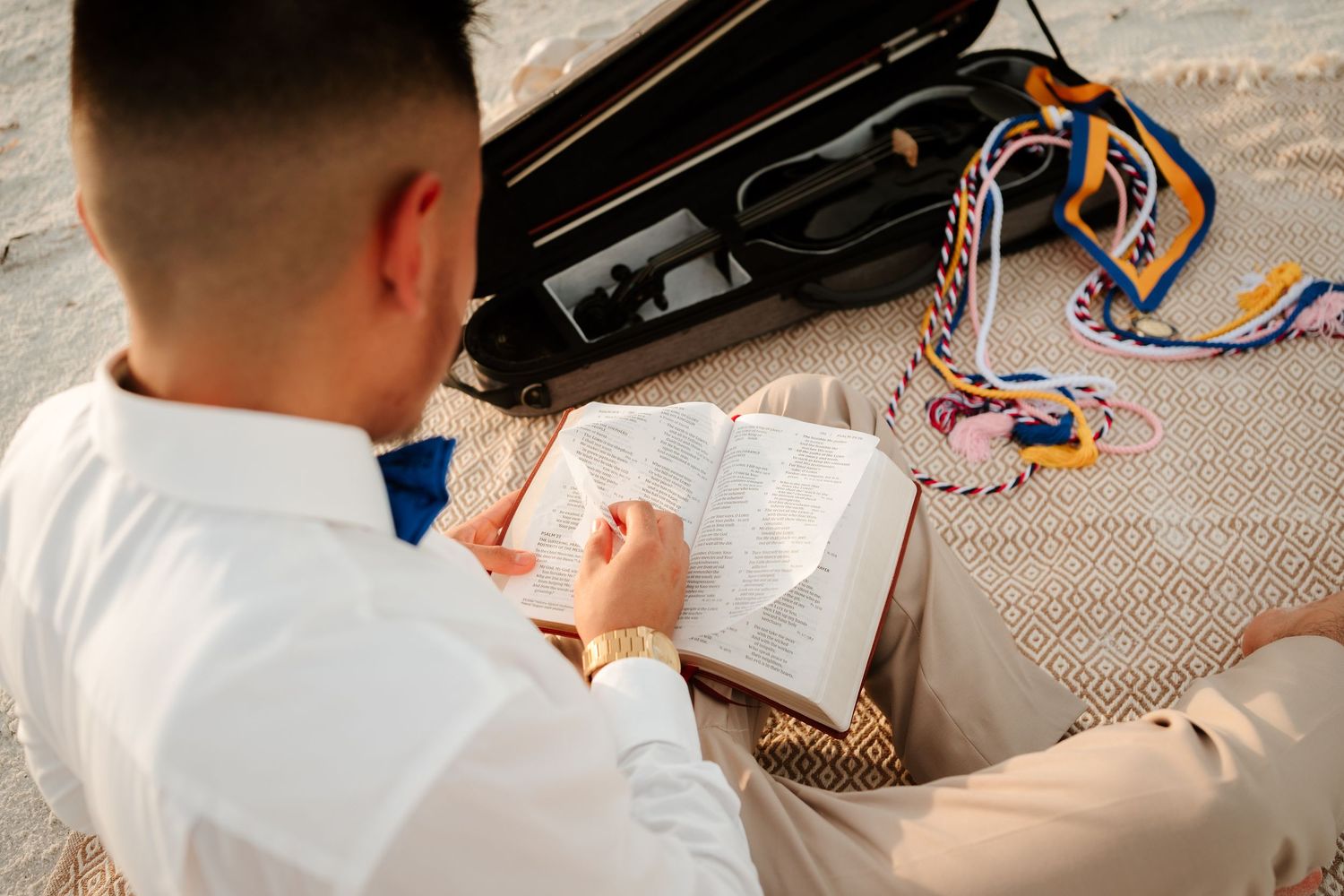 Man in white shirt reads open book outdoors near violin case and colorful graduation cords on sandy ground.