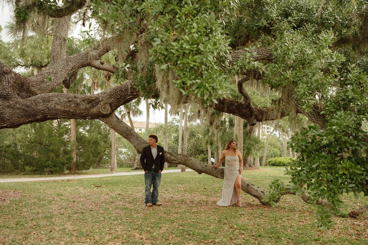 Bride and groom stand beneath a sprawling oak tree draped in Spanish moss in a lush green park setting.