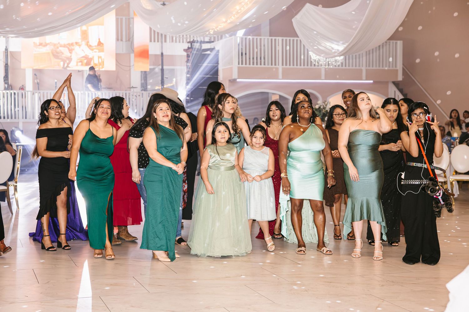 Diverse group of wedding guests in colorful formal attire gathered on dance floor during bouquet toss at reception.