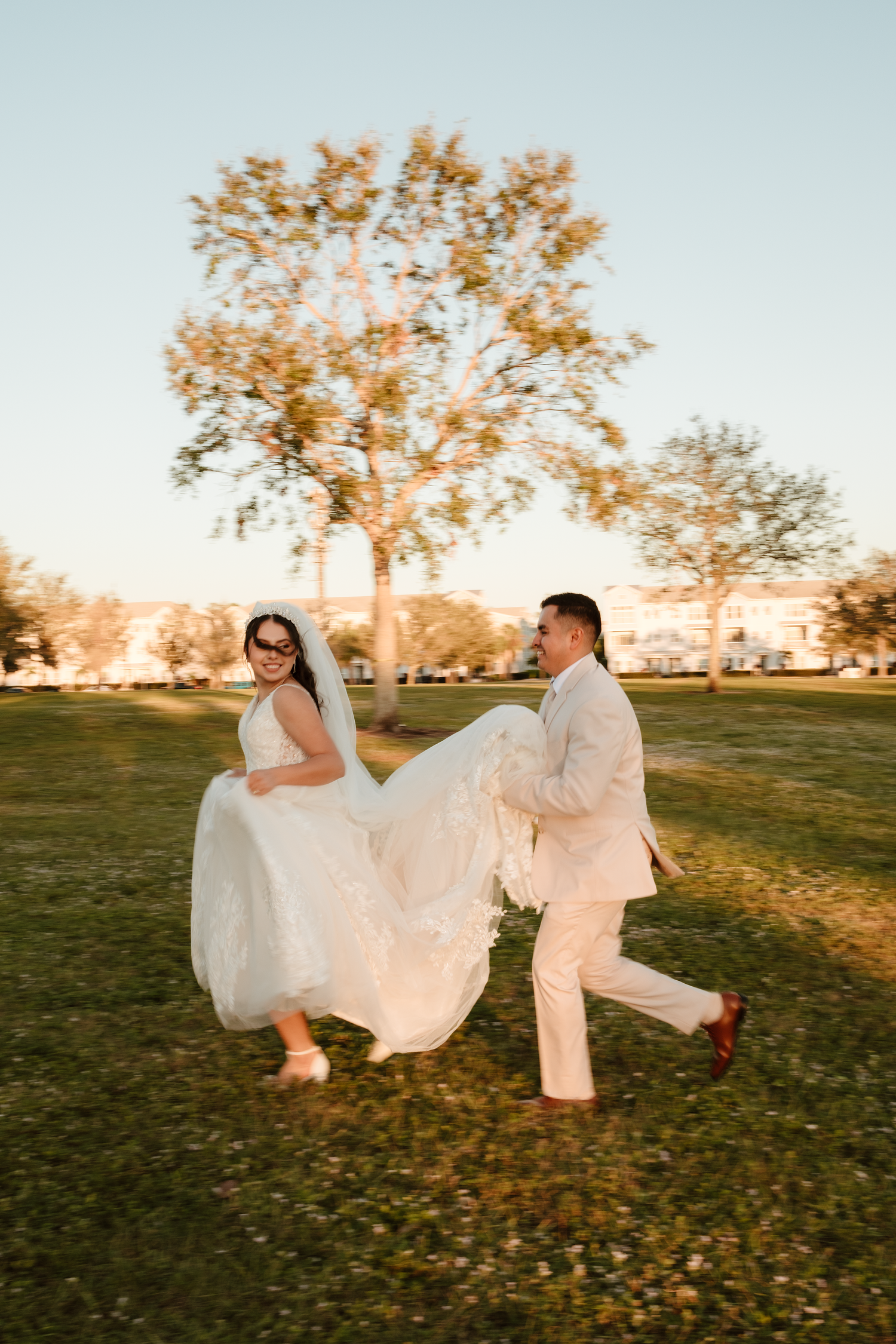 Joyful bride in white gown and groom in cream suit playing in golden hour sunlight at park.