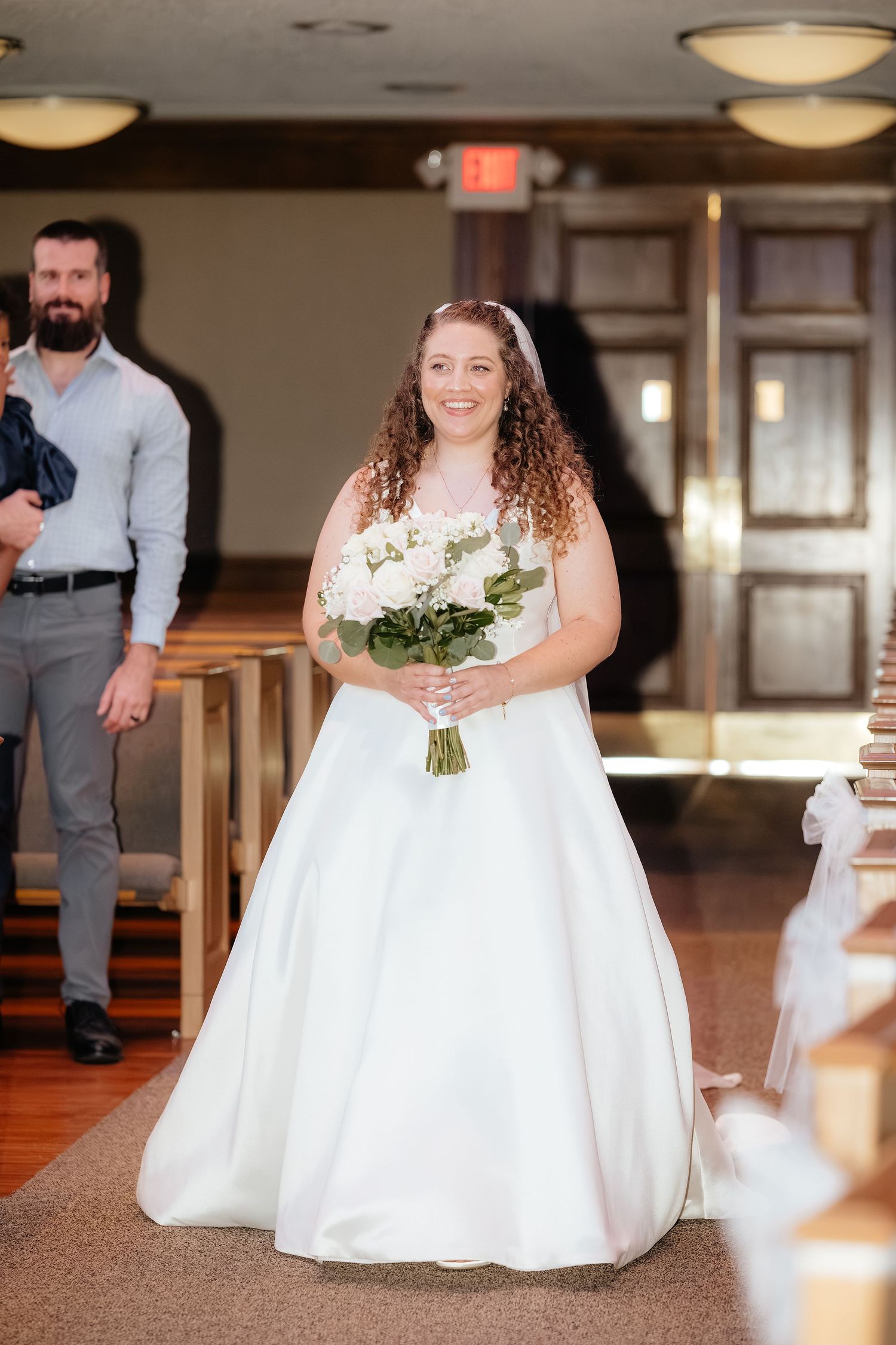 Smiling bride walking down church aisle in white ballgown holding floral bouquet.