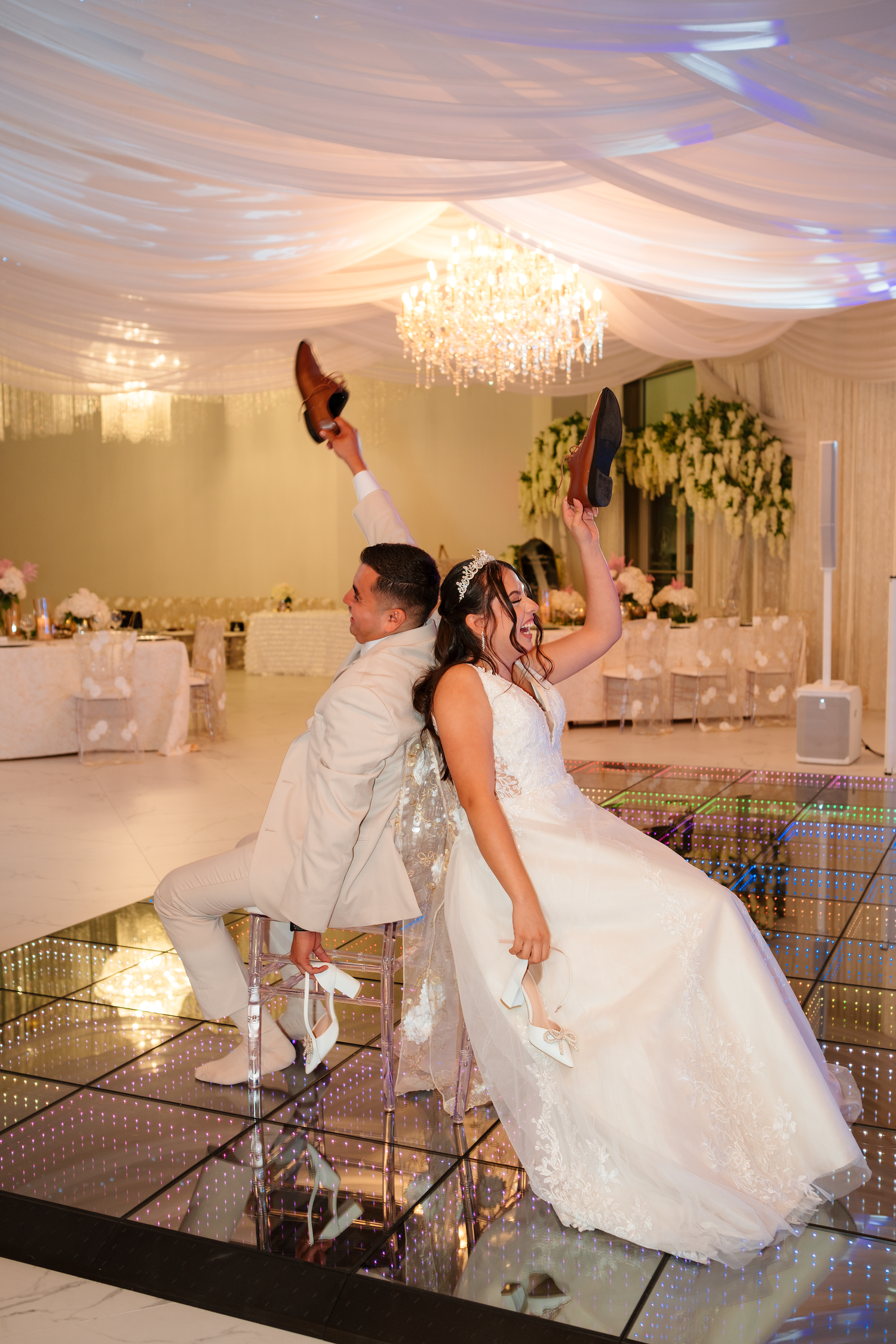 Bride and groom playing the shoe game seated on chairs on a mirrored dance floor.