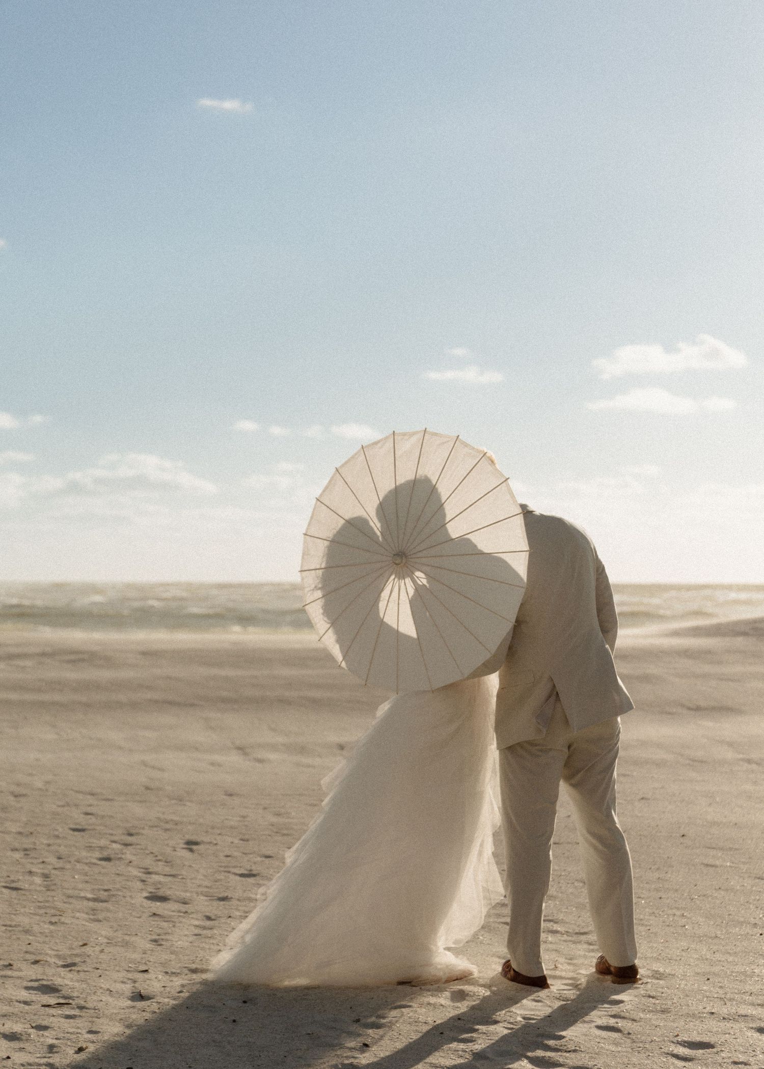 Beach wedding couple kissing behind white parasol umbrella, bride in gown and groom in tan suit.
