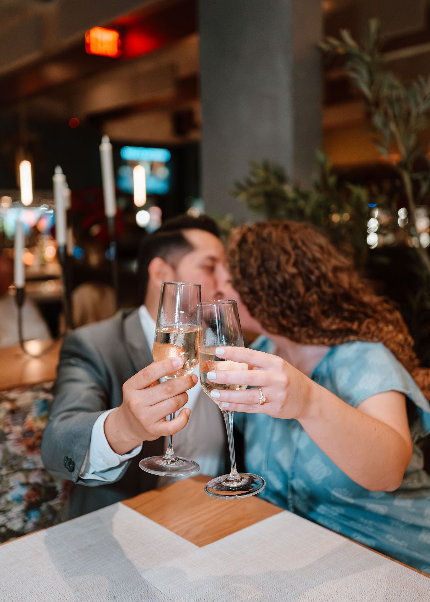 Couple kissing and toasting with champagne flutes at a restaurant table, celebrating a romantic occasion.