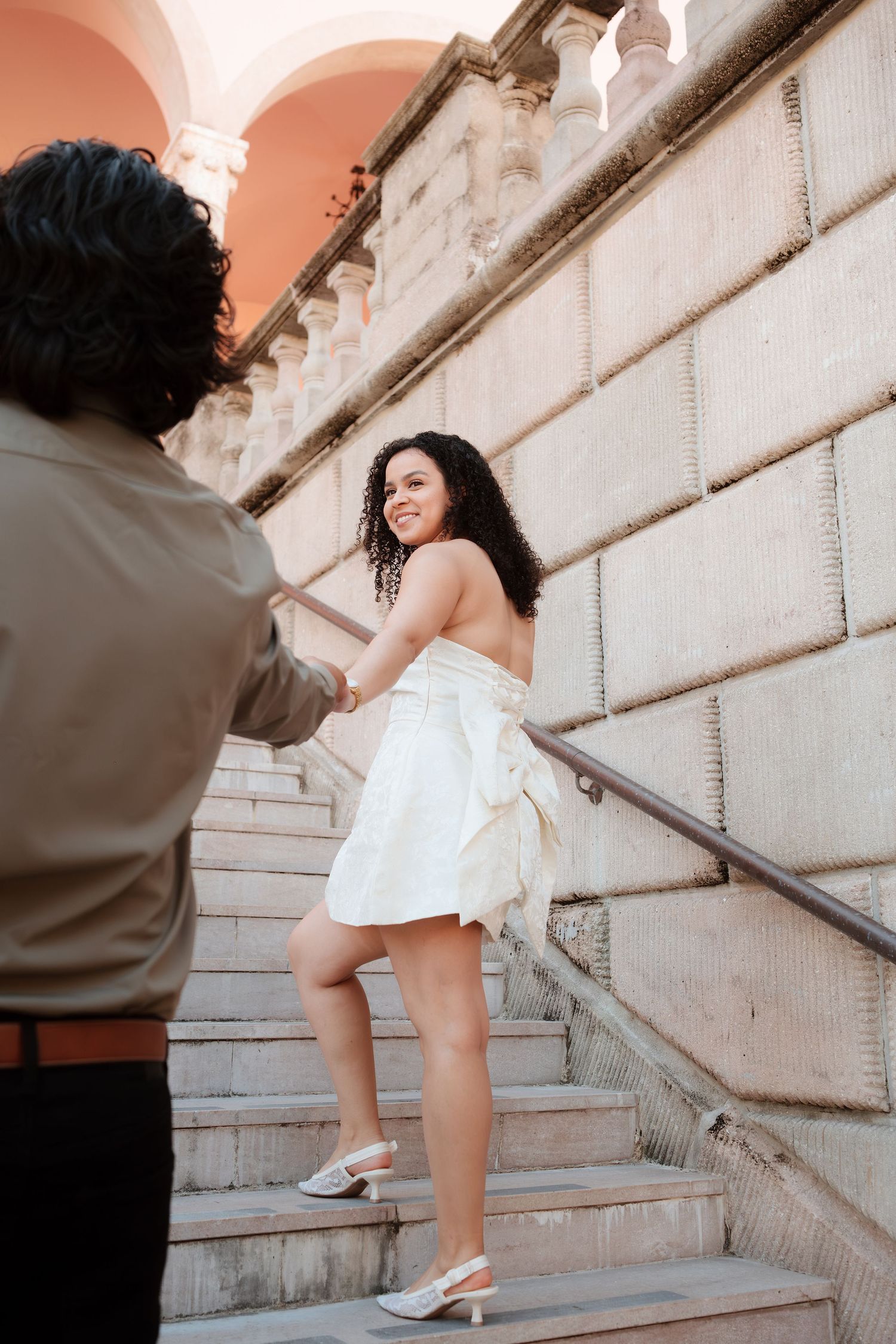 A smiling bride in a white dress holds hands with her partner while ascending elegant stone stairs.