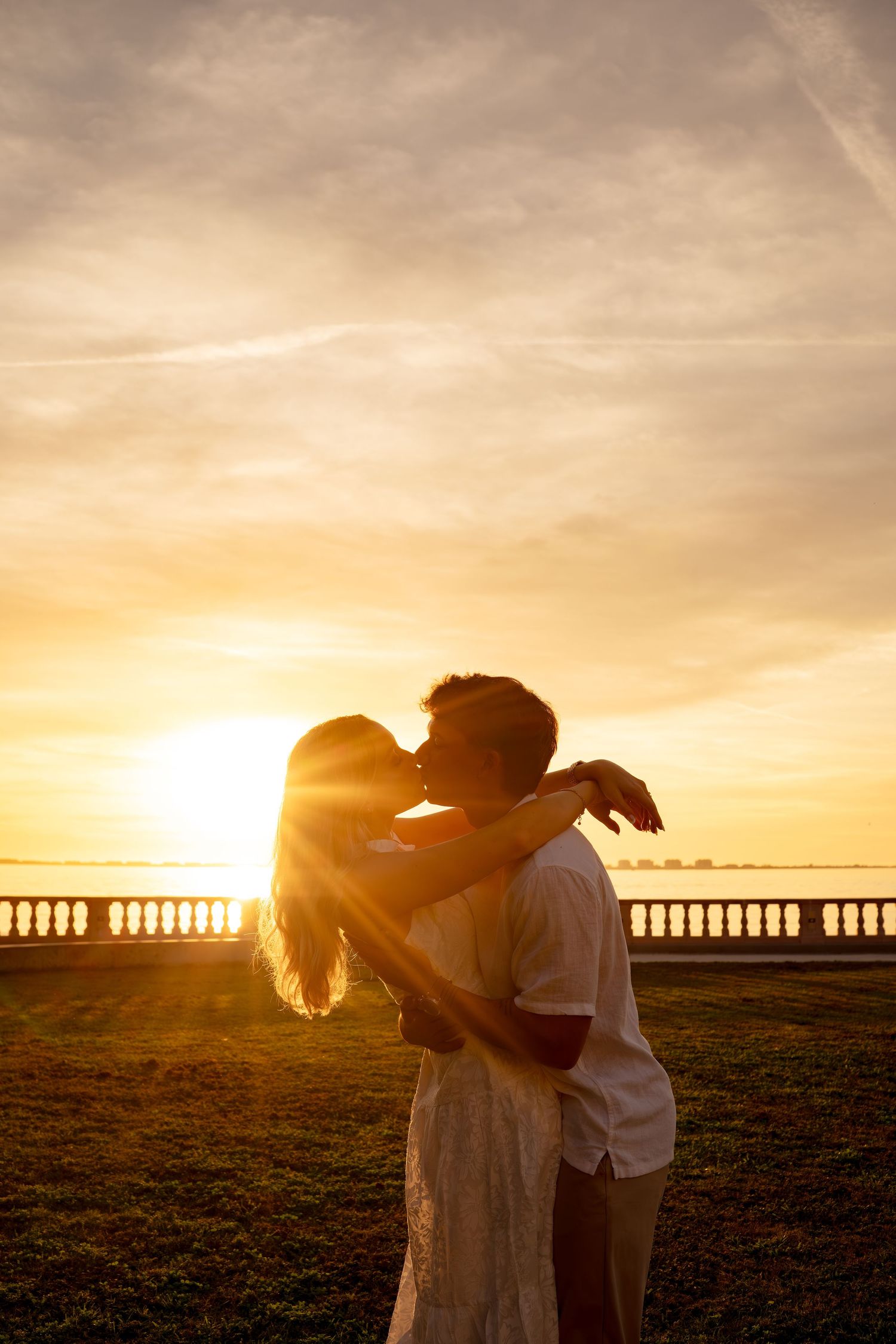 Romantic couple kissing at golden sunset by the waterfront, silhouetted against warm glowing sky.