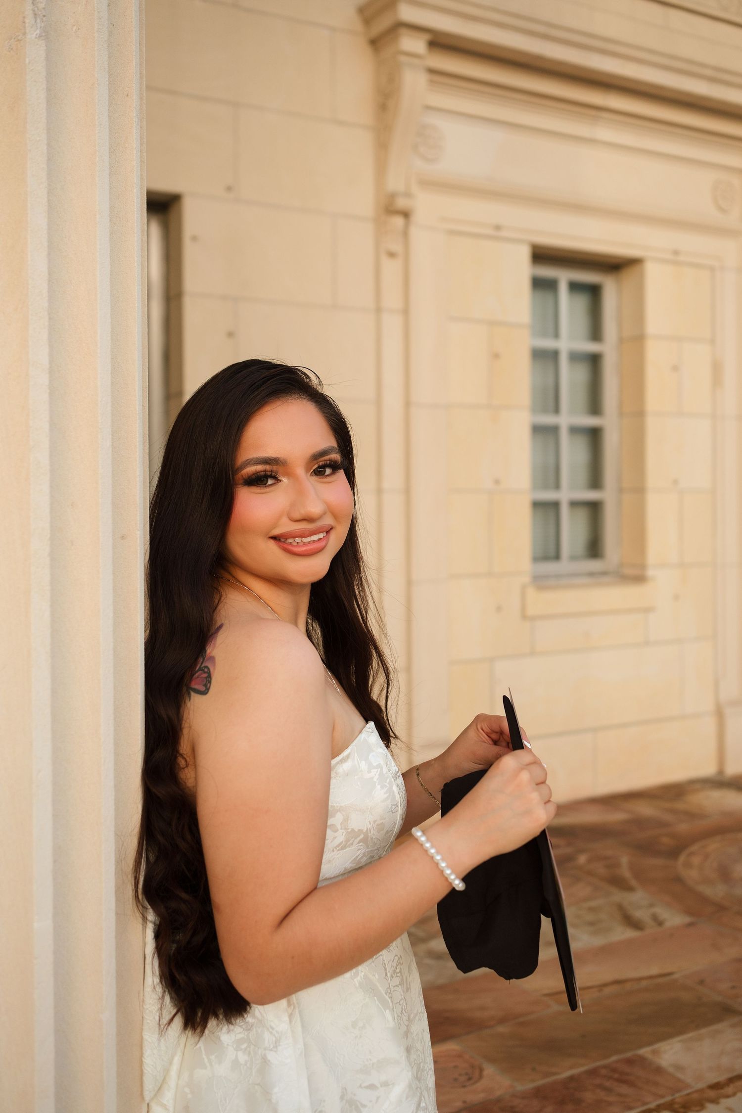 Young woman in white dress holding graduation cap, smiling beside a stone column at a university building.