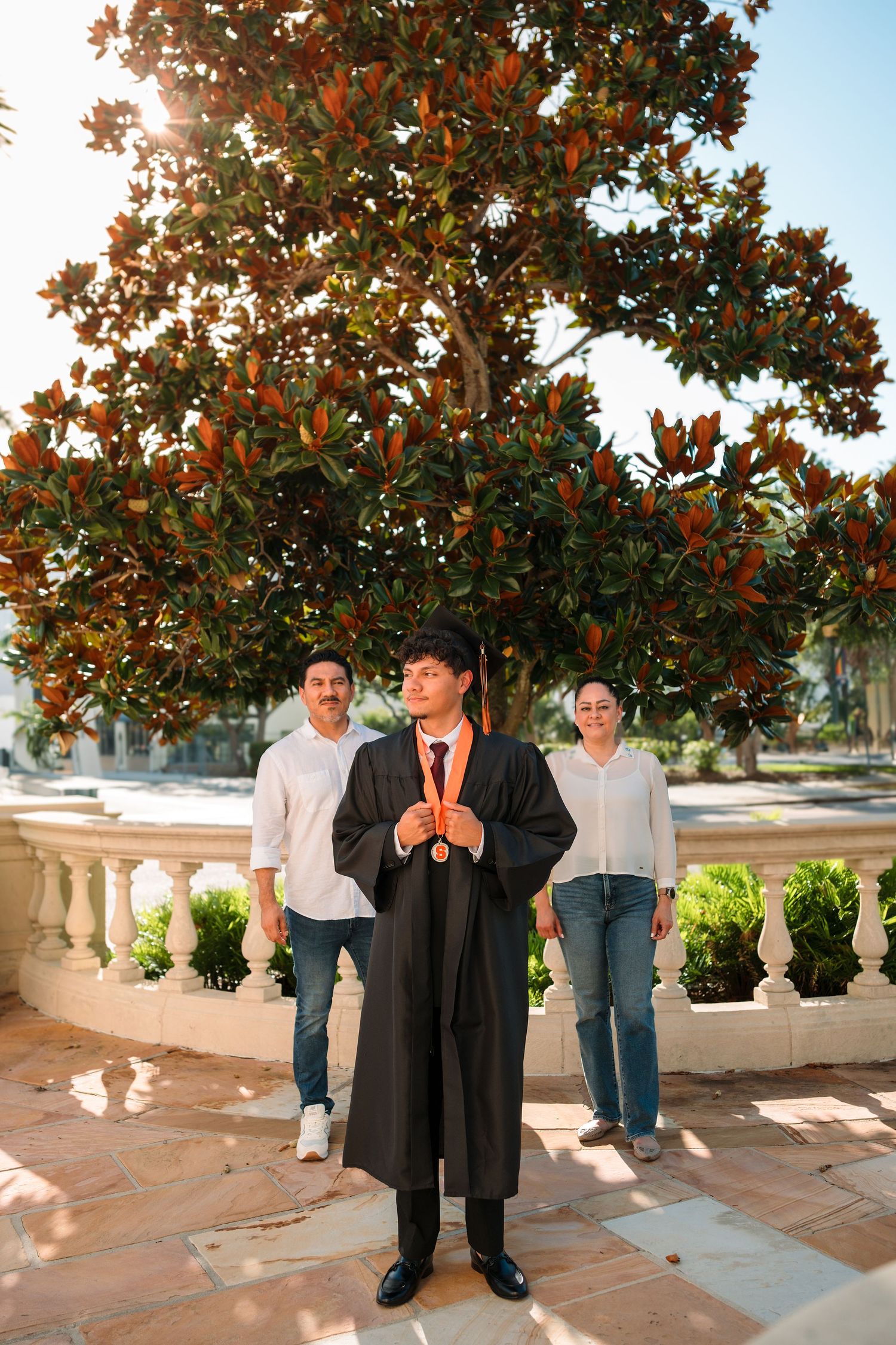 Graduate in cap and gown poses with proud parents before a magnolia tree on a sunny graduation day.