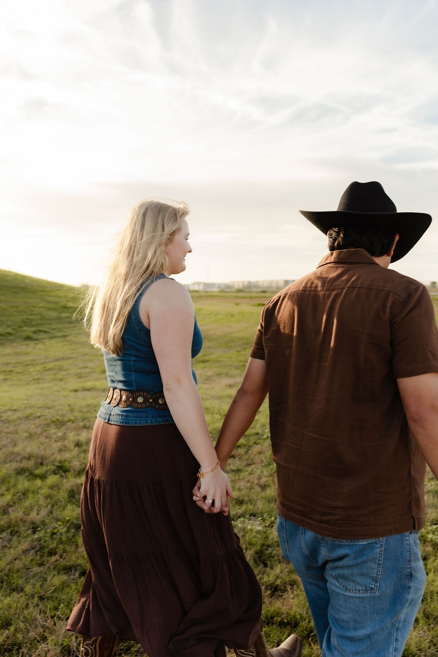 Couple holding hands walking through green pasture at golden hour sunset.