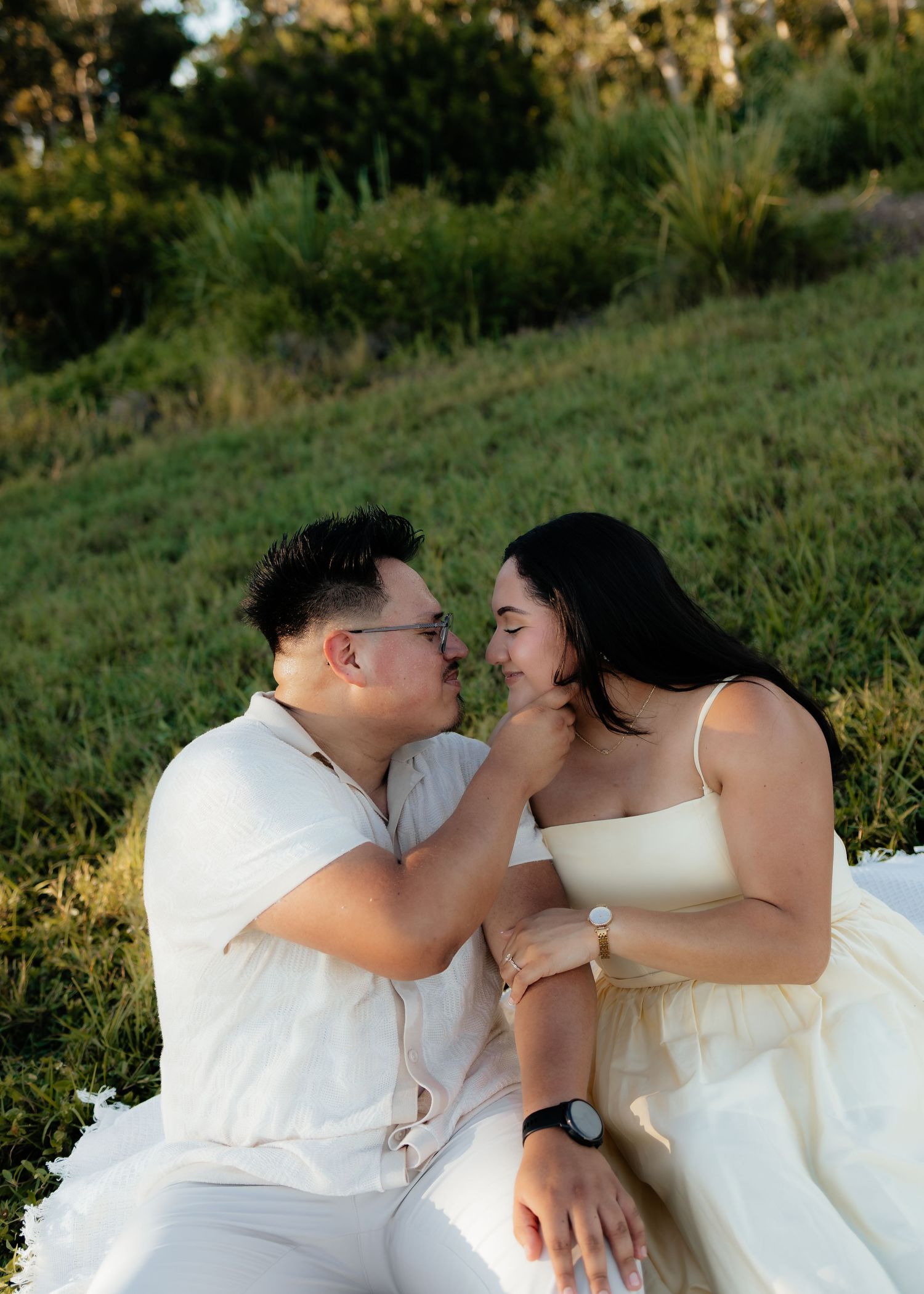 Couple in white outfits sitting on grass, sharing a tender moment at golden hour during an outdoor engagement session.
