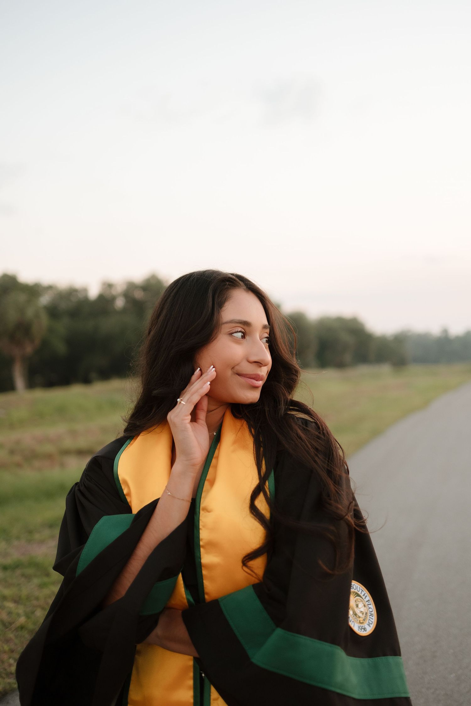 Young woman in black and gold graduation regalia smiling outdoors on a road surrounded by green fields at dusk.