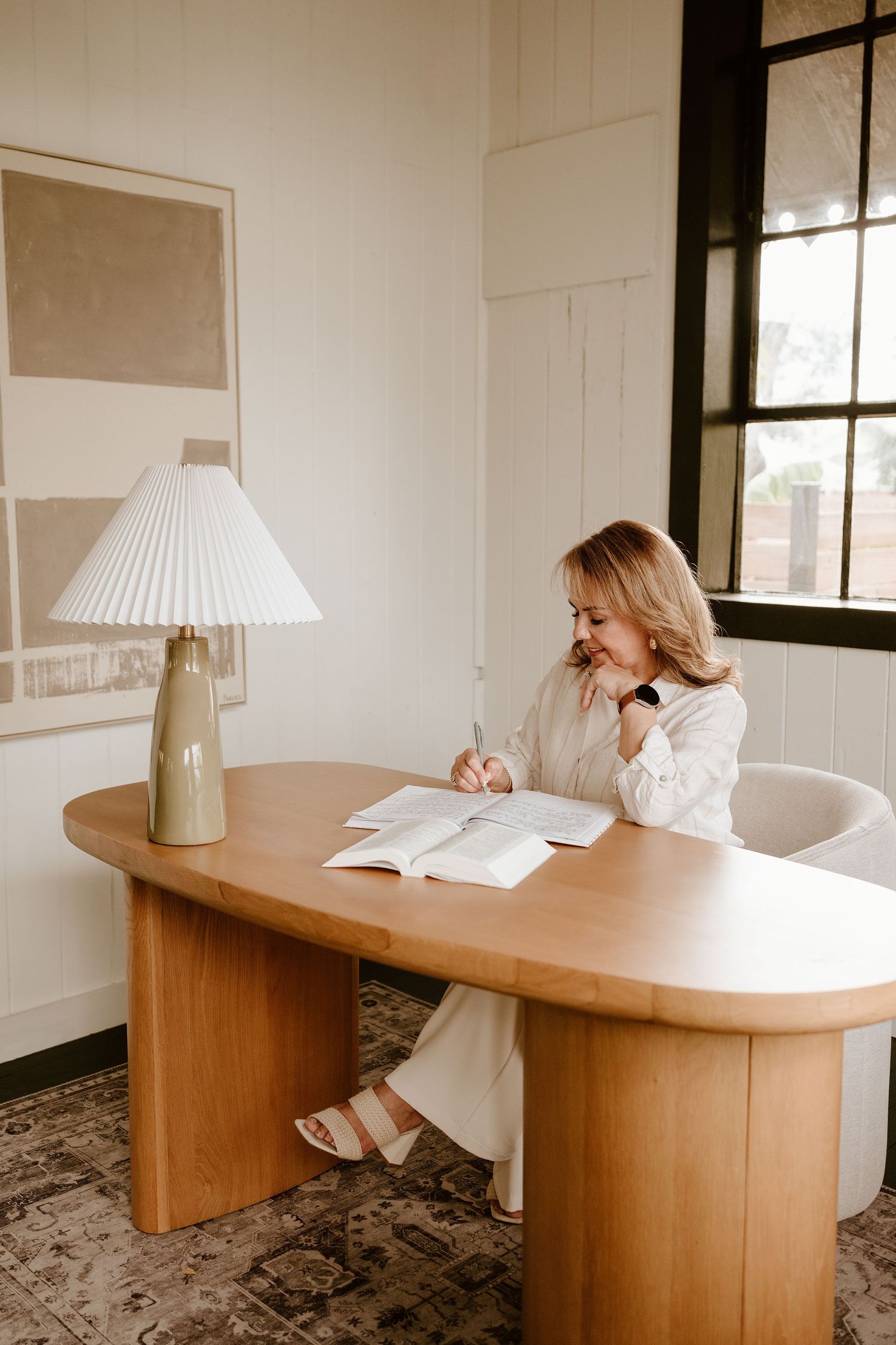 Woman in white outfit writing in open book at oval wooden desk beside lamp and window in bright room.