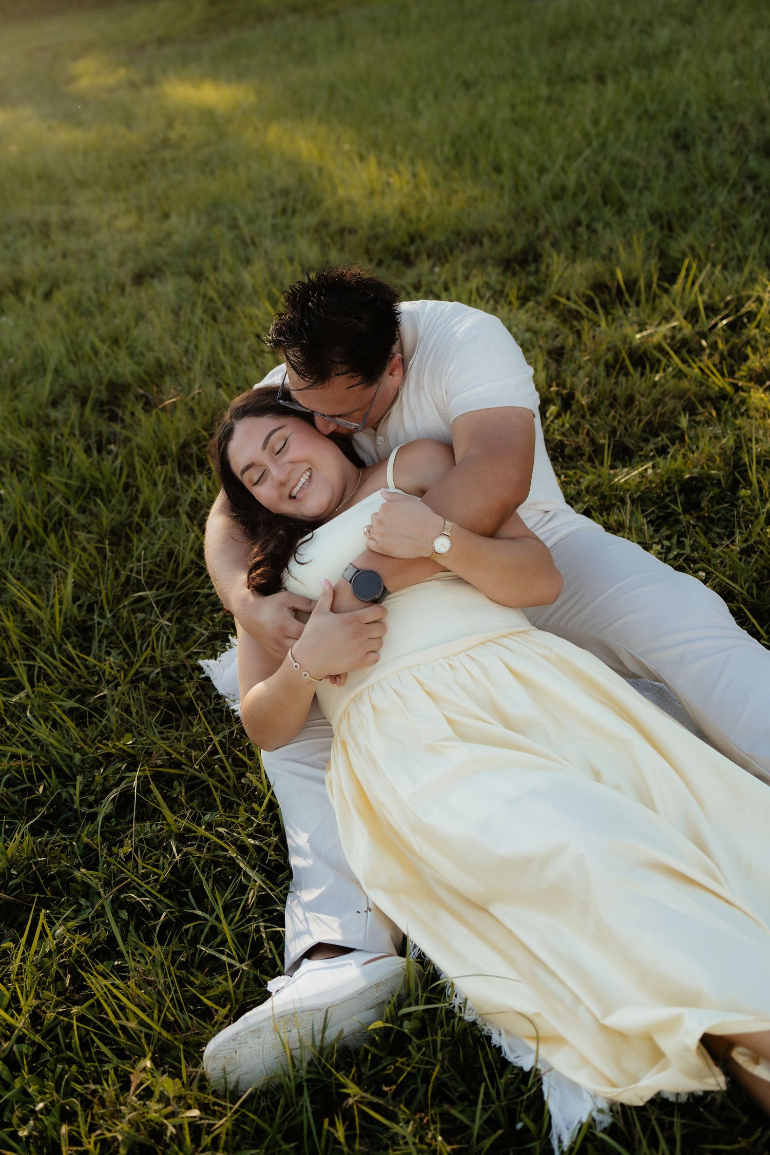 A joyful couple relaxing on green grass, woman in yellow dress laughing as man in white embraces her.
