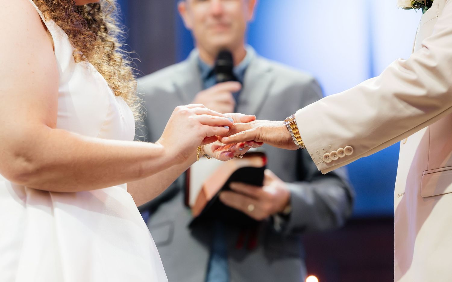 Bride placing wedding ring on groom's finger during ceremony, with officiant holding microphone in background.