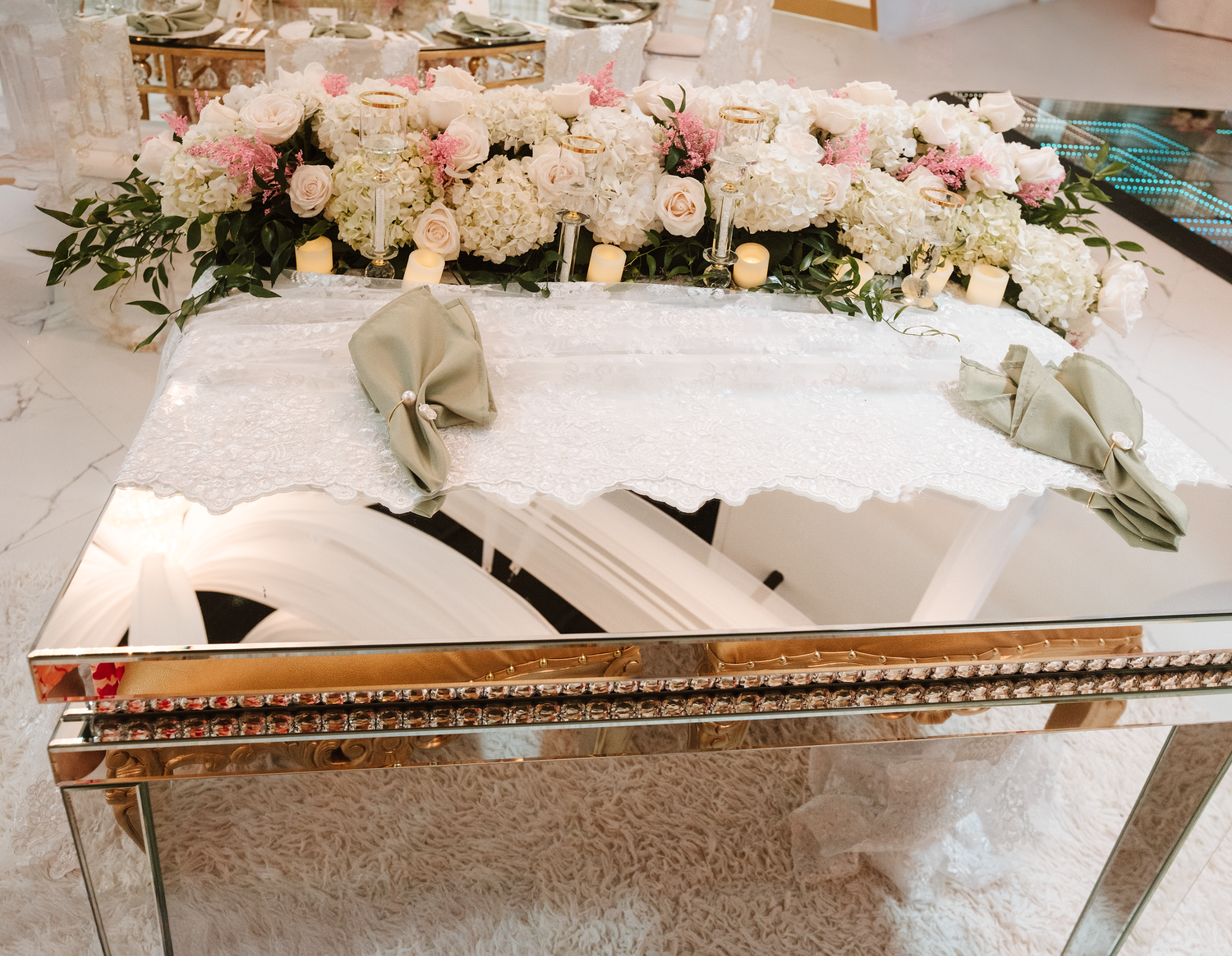 Elegant wedding reception table with lush white and pink floral centerpiece, mirrored gold table, and sage green napkins.