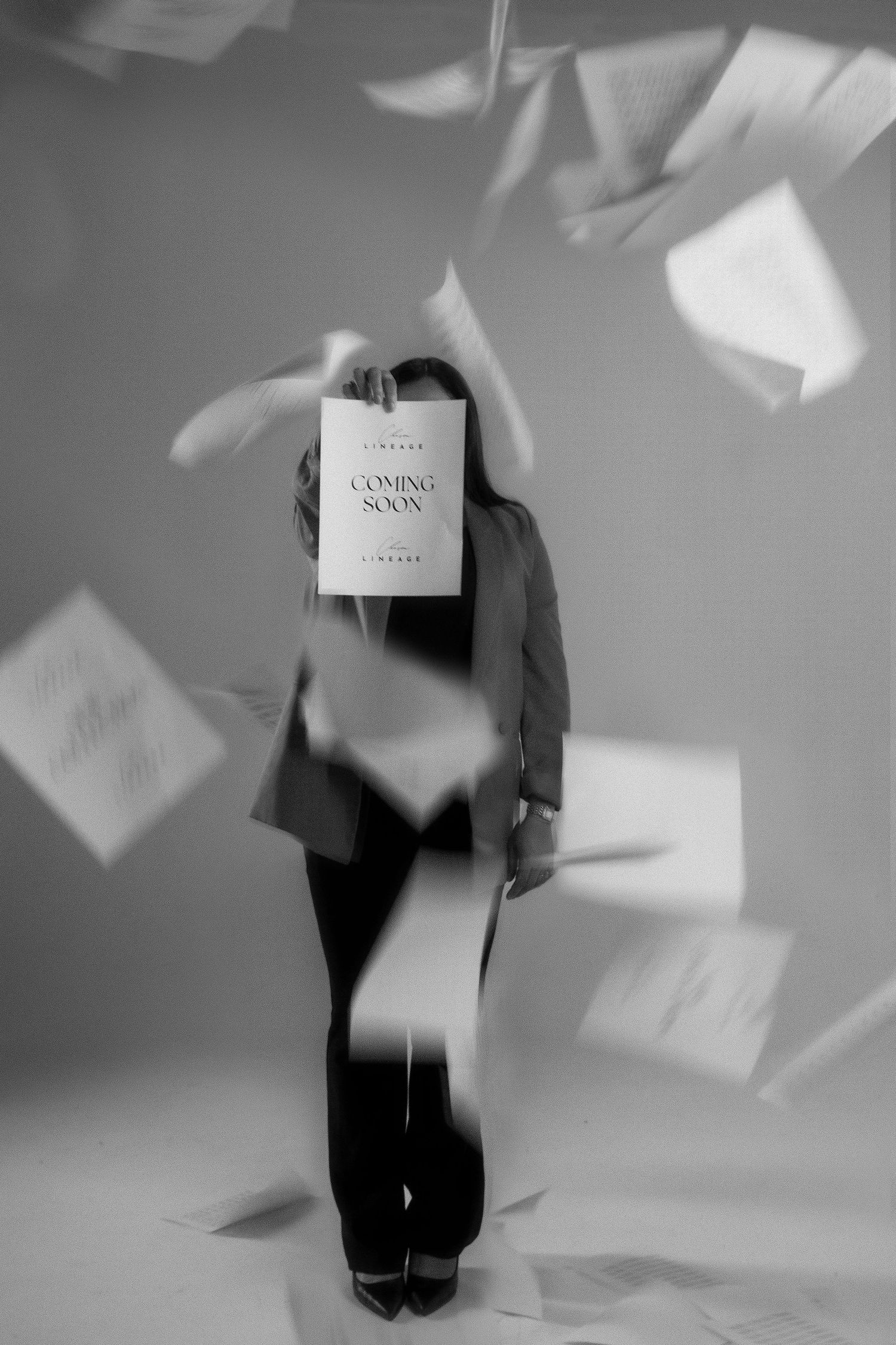 Black and white photo of person holding Coming Soon sign with papers flying around them in studio setting.