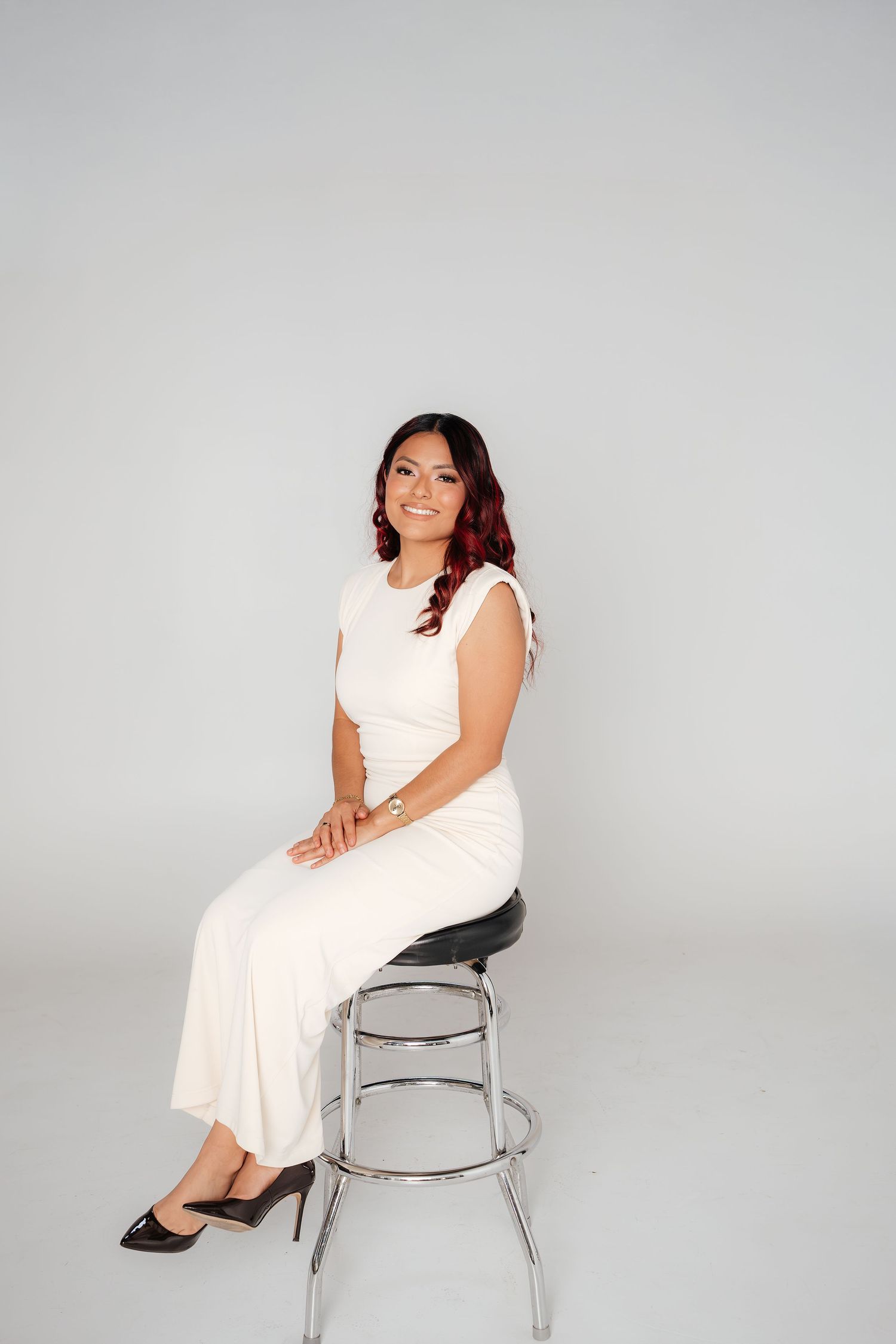 A woman in a white sleeveless dress sits gracefully on a bar stool against a light gray studio background.