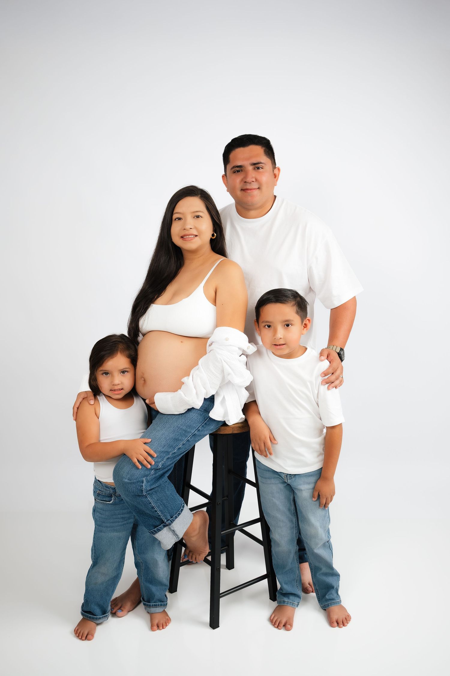 Pregnant Hispanic family posing in white tops and jeans for a maternity photo shoot with two young children.