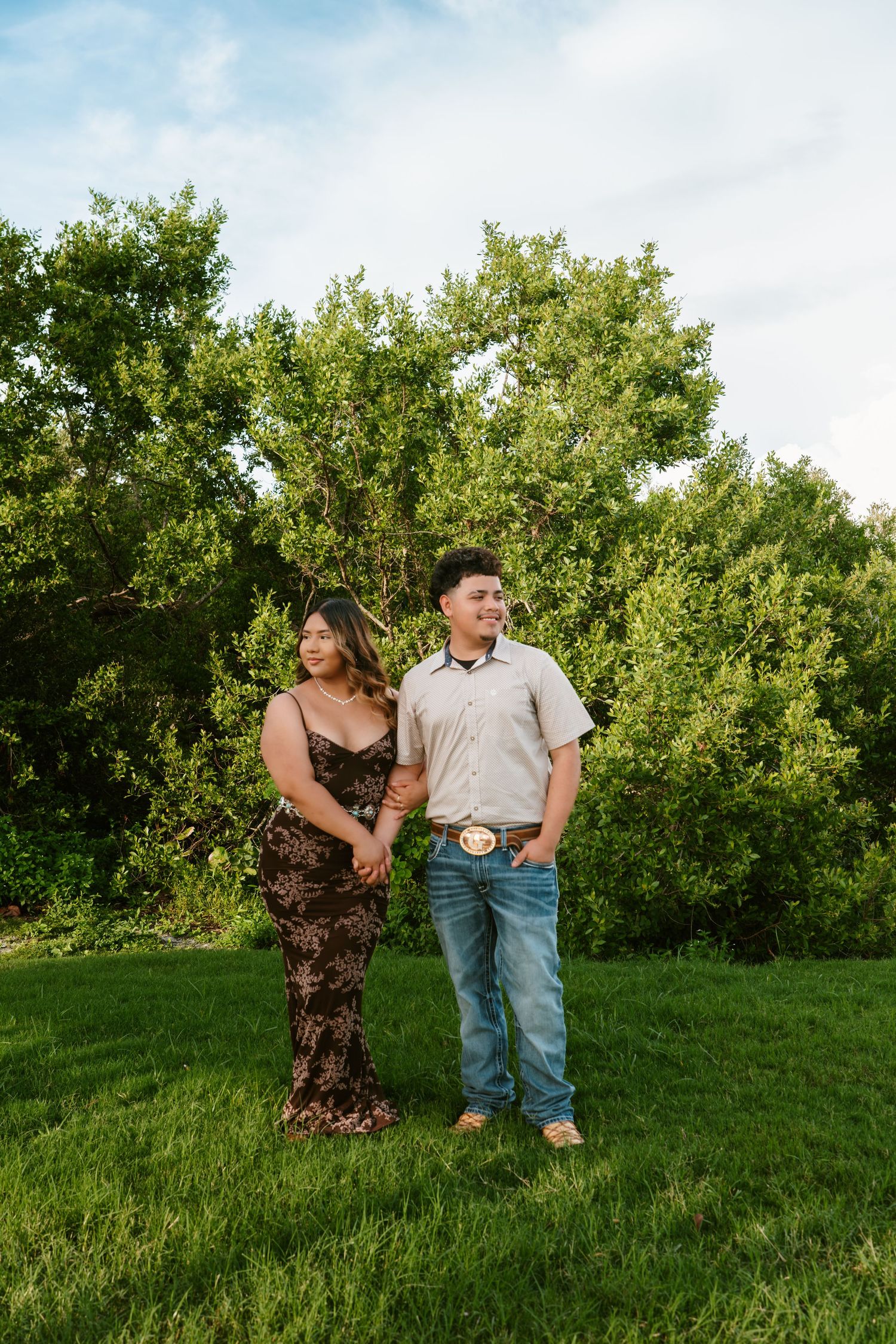 A couple holds hands on green grass, woman in floral dress and man in jeans with belt buckle, trees behind them.