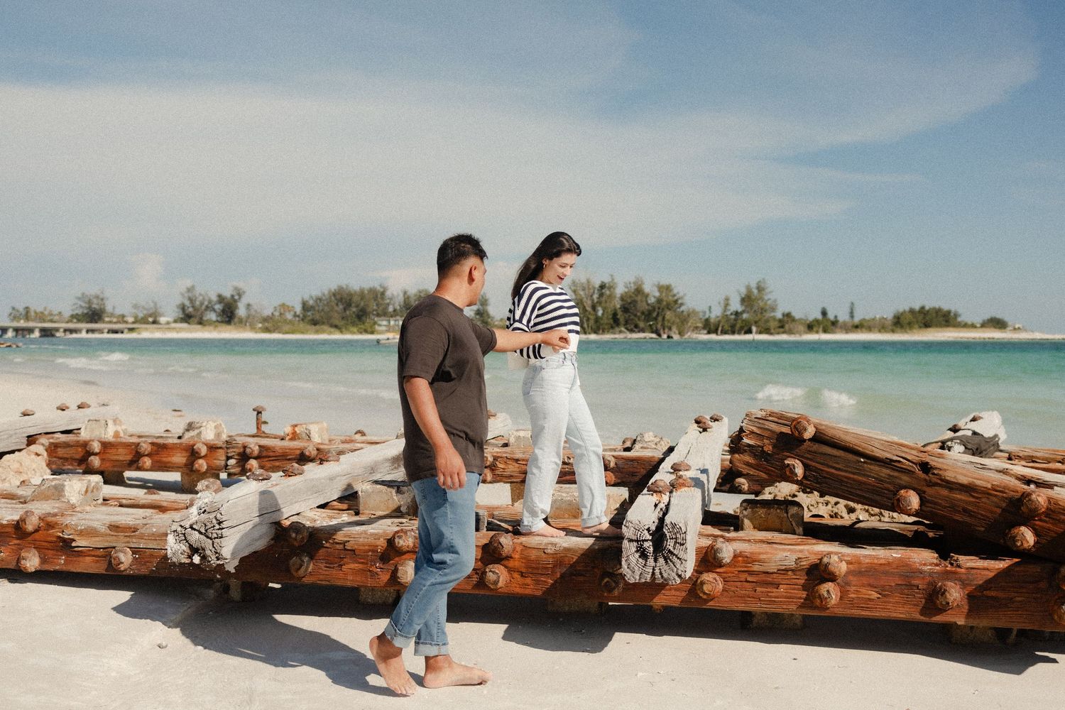 Couple walking along a sandy beach with driftwood logs, turquoise water and trees in the background on a sunny day.