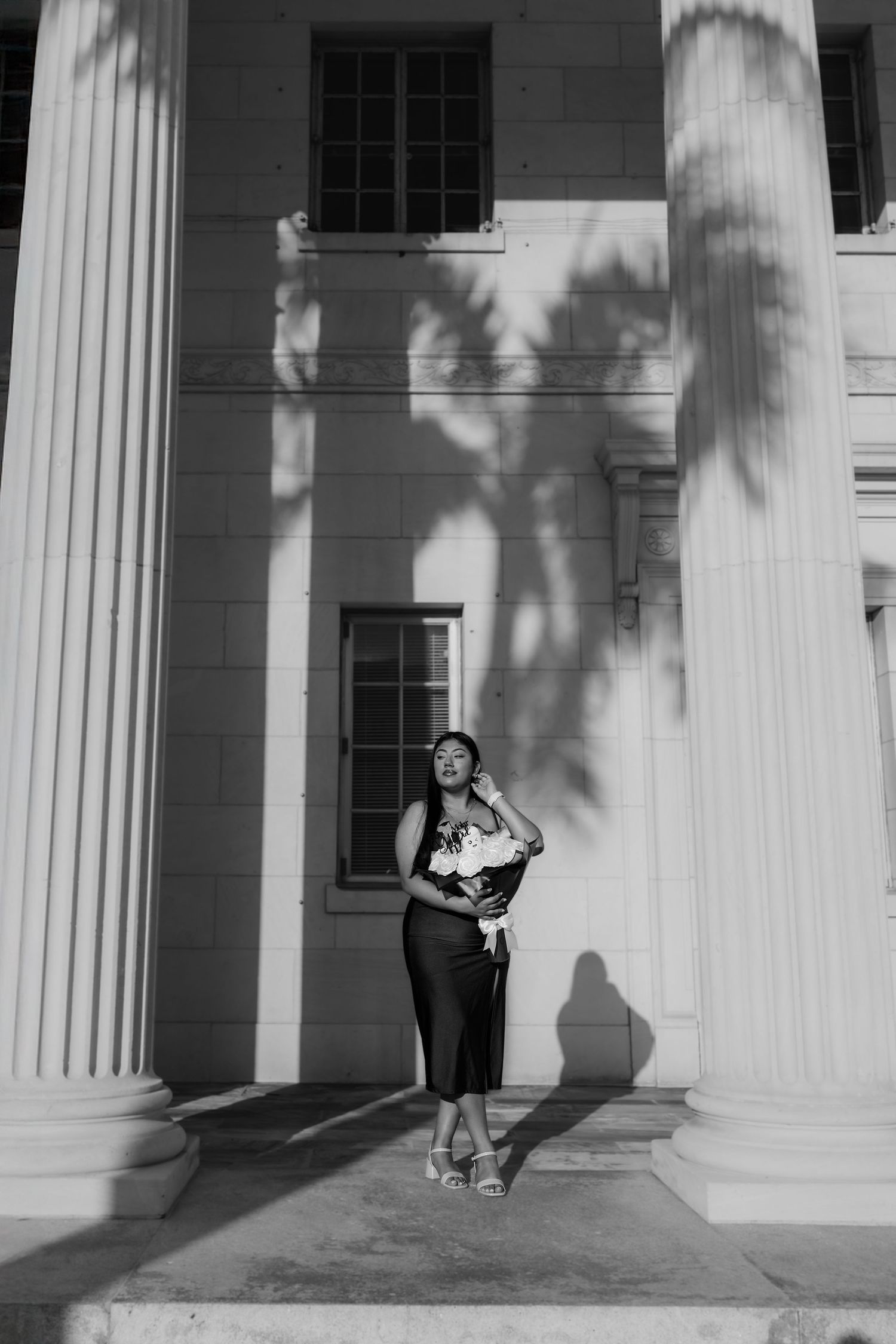 Black and white photo of a woman in a midi dress holding flowers, posing between large columns of a classic building.