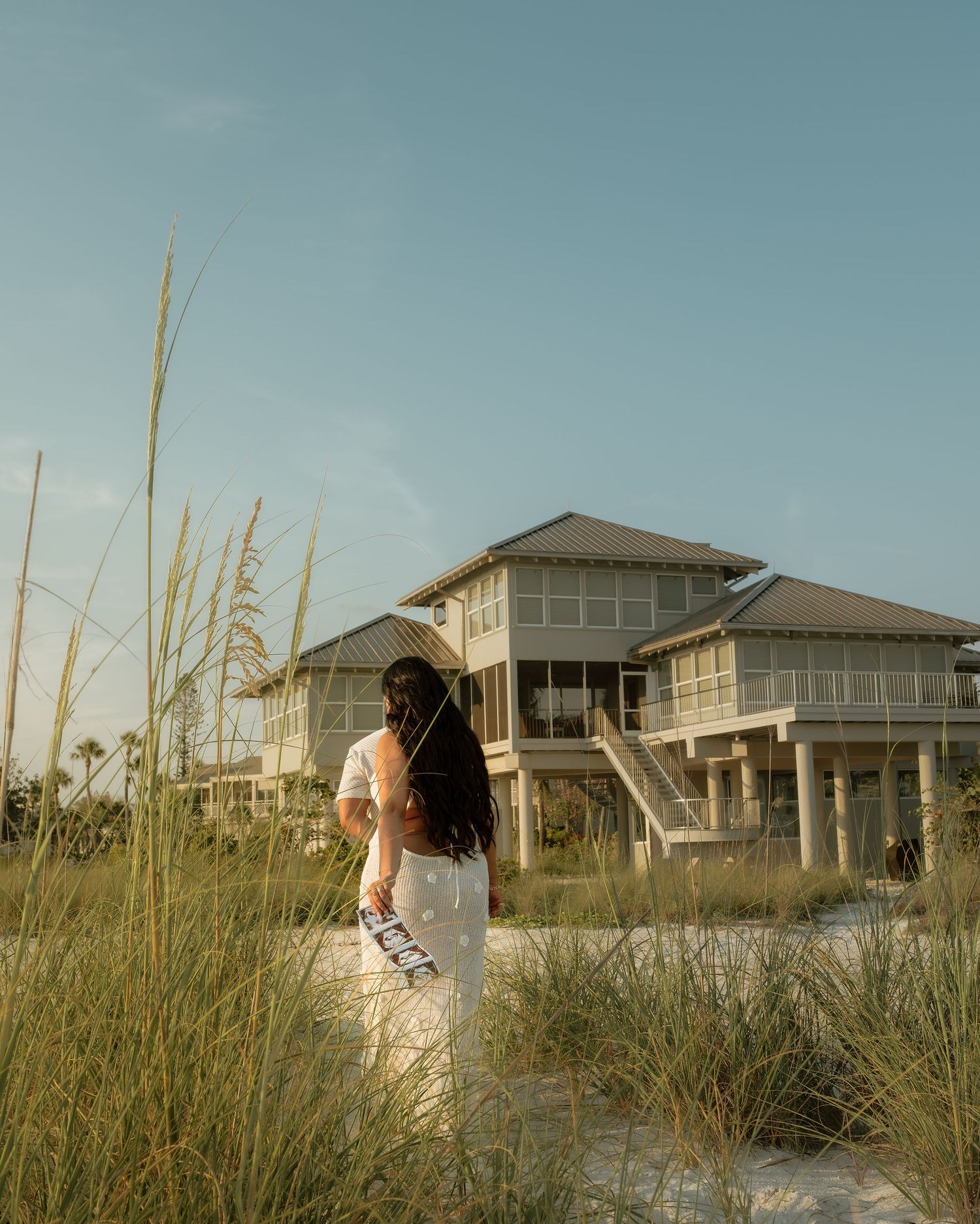 A bride in a white gown stands on a sandy beach path surrounded by sea grass, facing a raised coastal house.