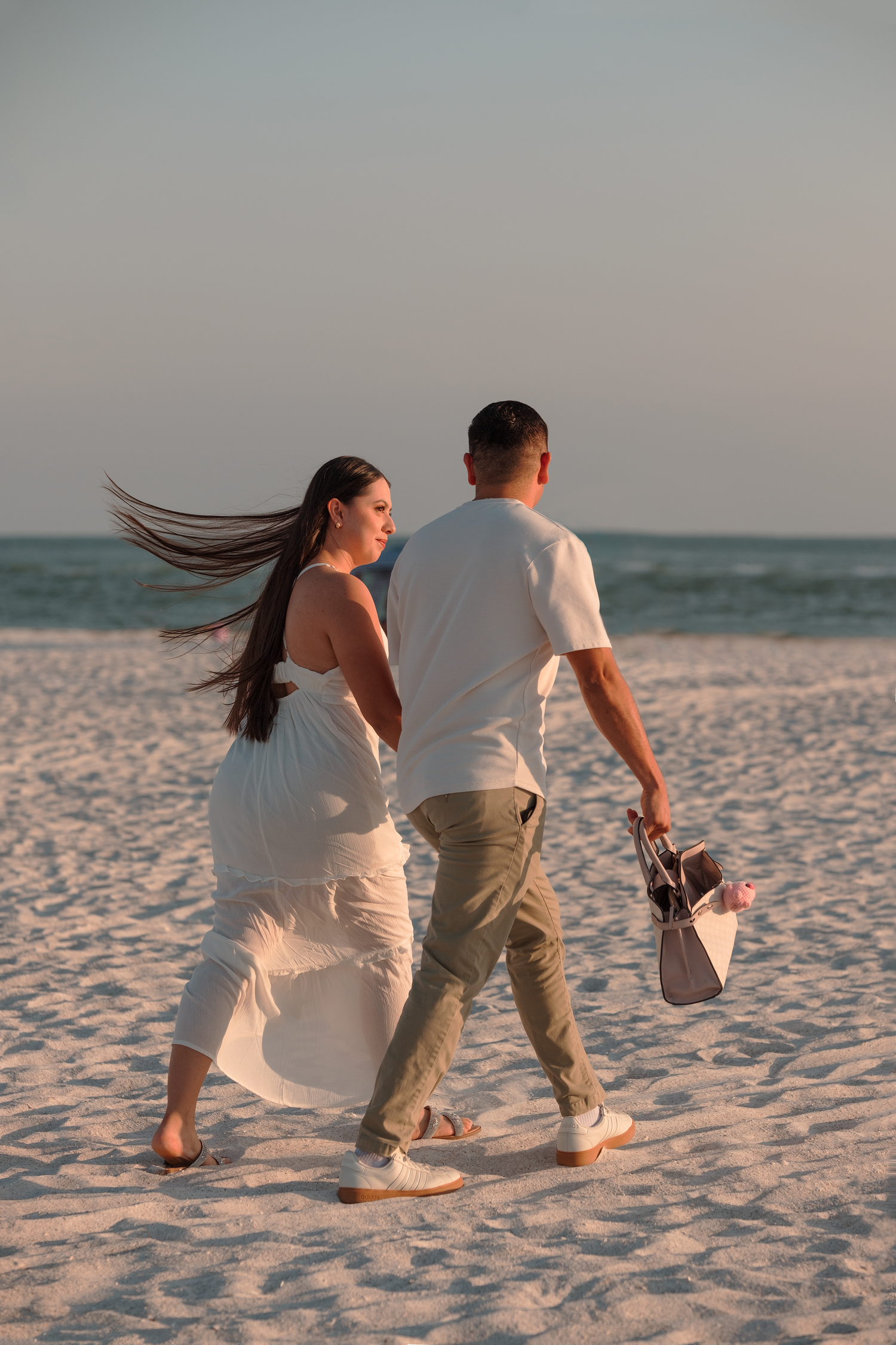 Couple walking on sandy beach at sunset, woman in white dress with flowing hair, man carrying shoes.