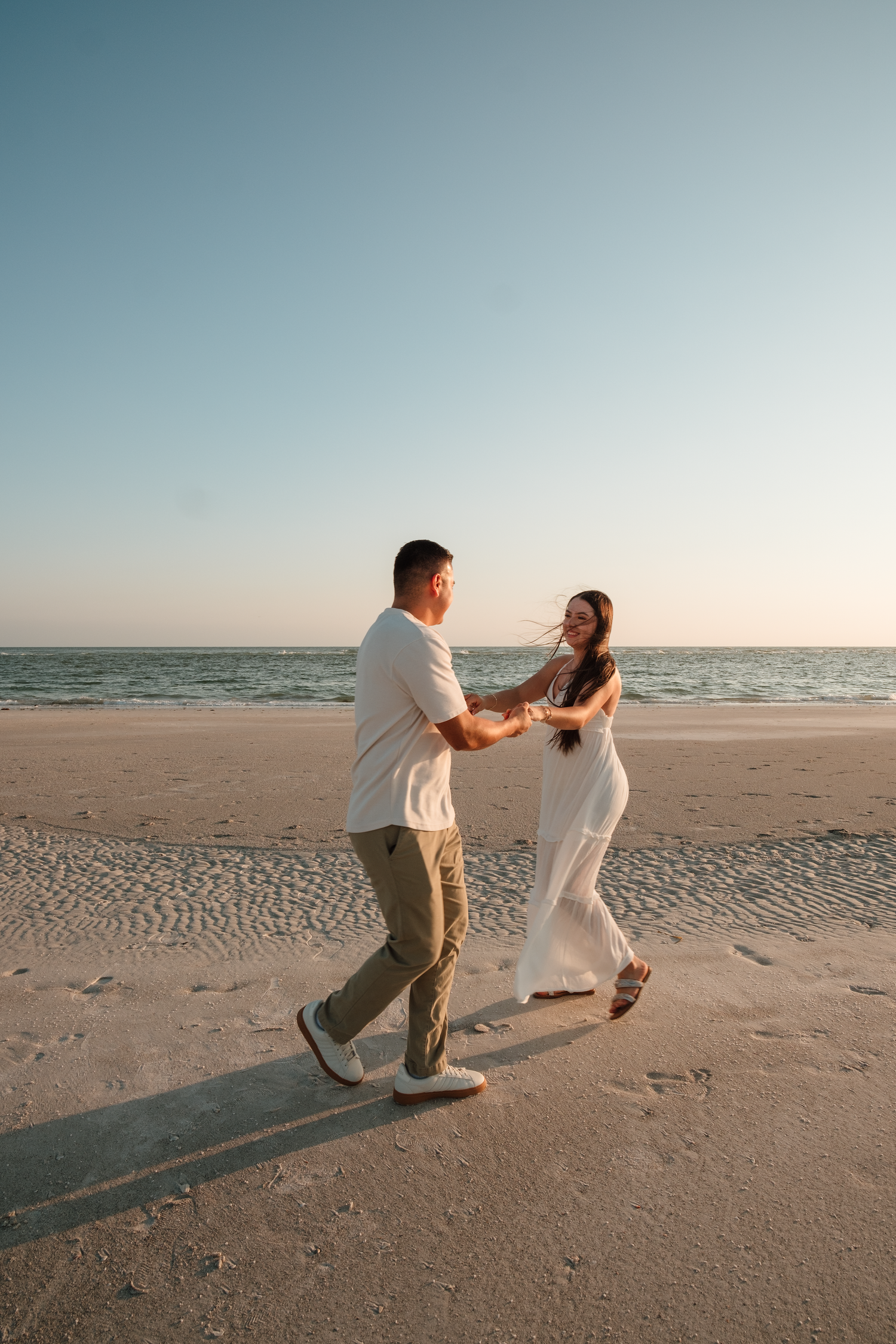 Couple dancing joyfully on a sandy beach at sunset, holding hands in white and neutral outfits with ocean in background.