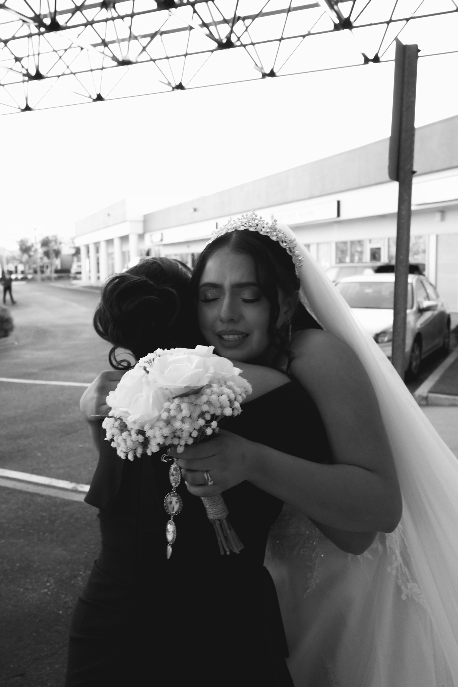 Black and white photo of a bride in veil and tiara embracing a guest while holding a white rose bouquet outdoors.