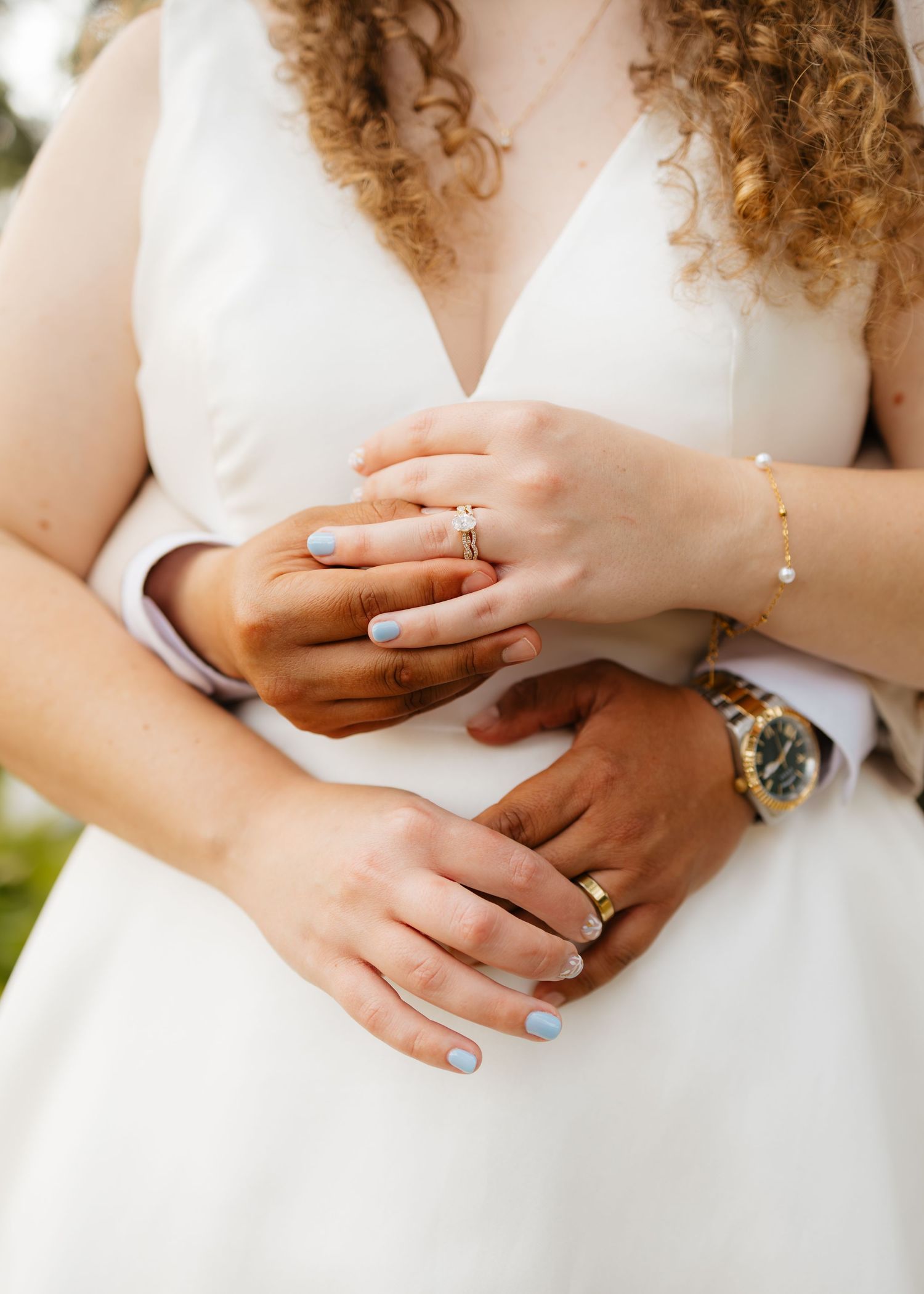 Close-up of bride and groom holding hands showing wedding rings, blue nails, and gold bracelet in white dress.