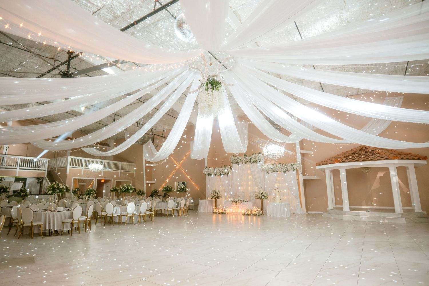 Elegant wedding reception hall with white draped ceiling fabric, round tables, floral centerpieces, and a spacious dance floor.