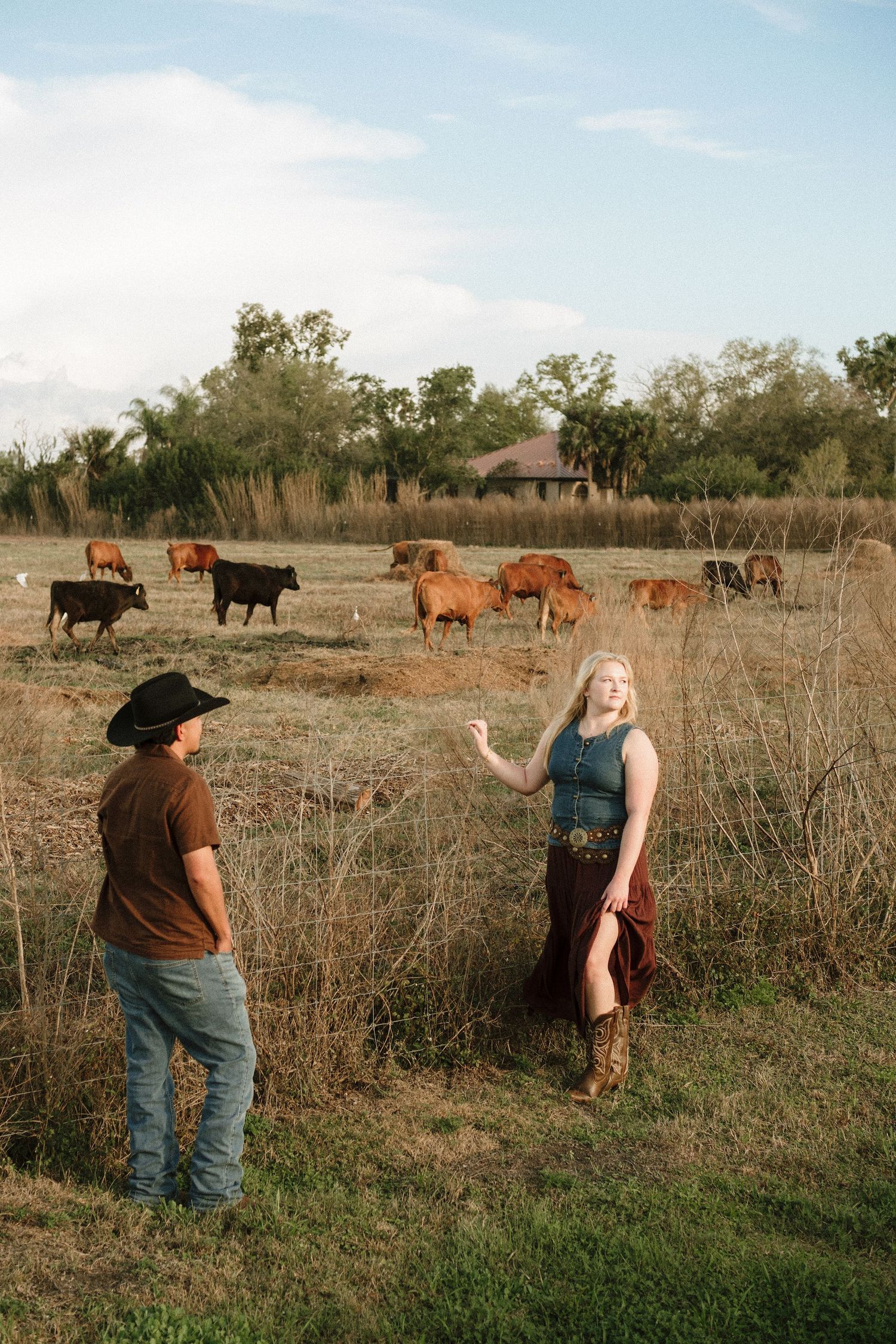 Two people in western wear converse in a rural field with grazing cattle and farmhouse in background.