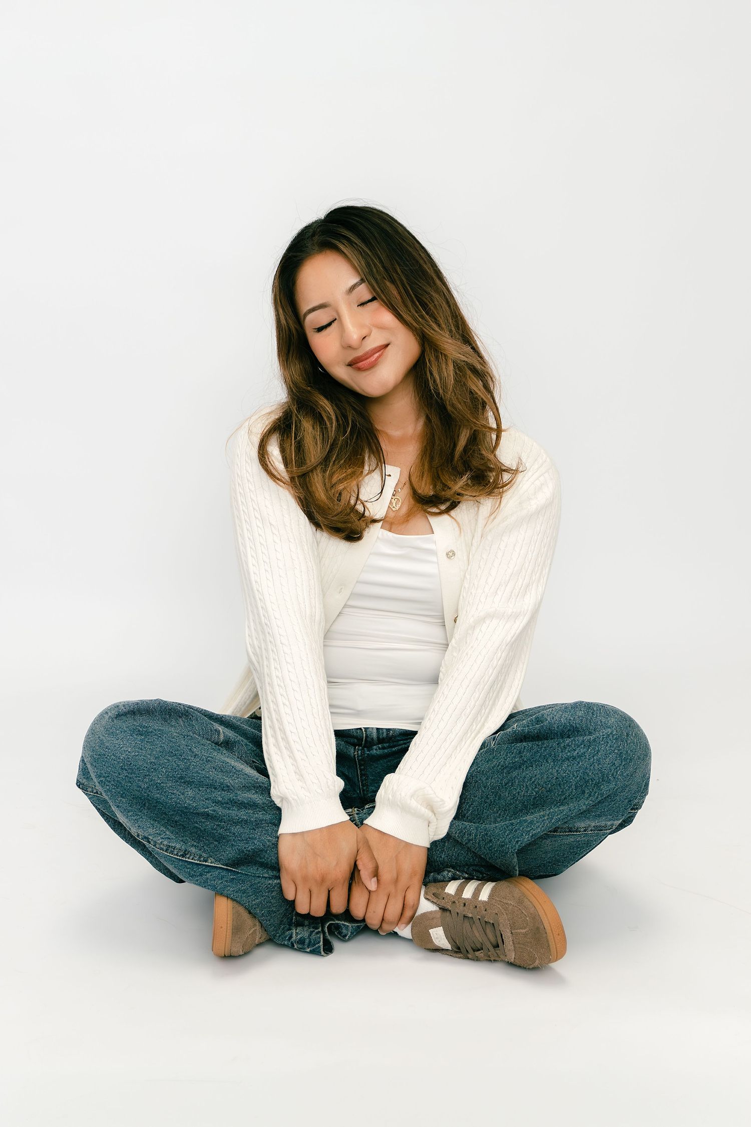 Young woman sitting cross-legged on white background, wearing white cardigan, jeans, and sneakers, smiling with eyes closed.