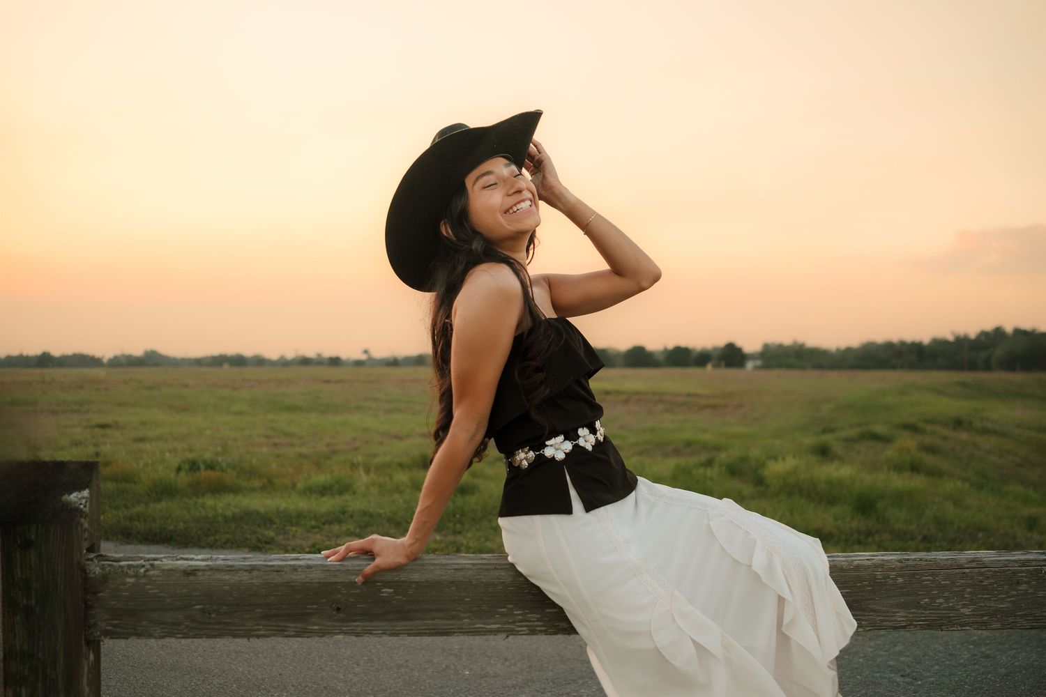 Woman in black cowboy hat and white skirt posing against fence at sunset in rural field.