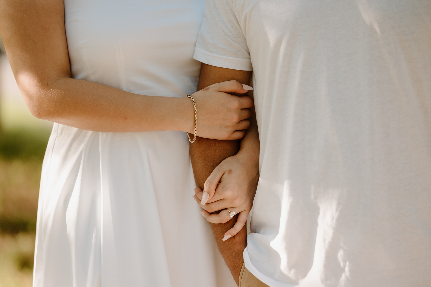A couple in white wedding attire holds hands while standing in a sunlit outdoor setting.