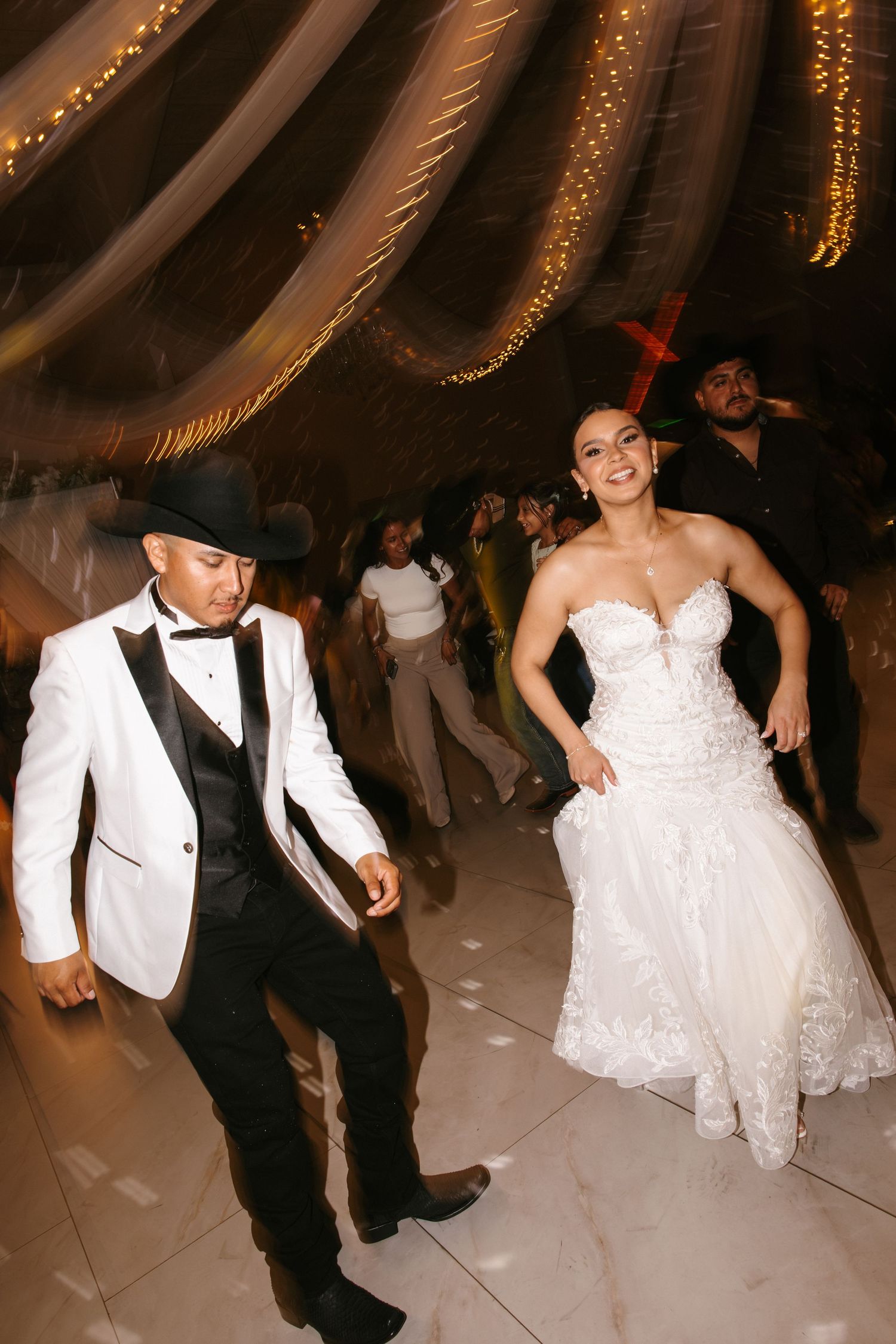 Bride in white gown and groom in white tuxedo with black cowboy hat dancing joyfully at wedding reception.