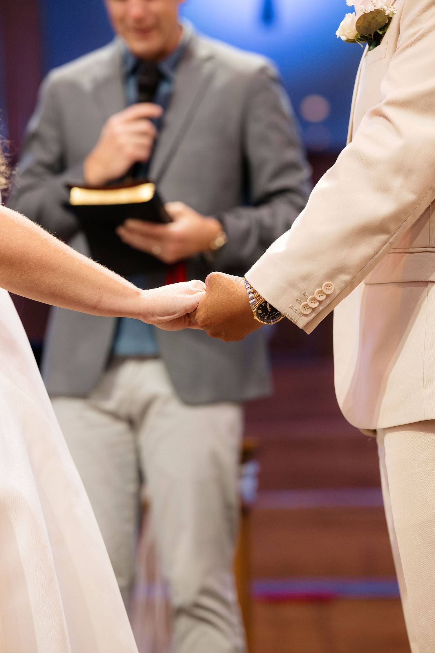 Bride and groom holding hands during wedding ceremony while officiant reads from Bible in background.