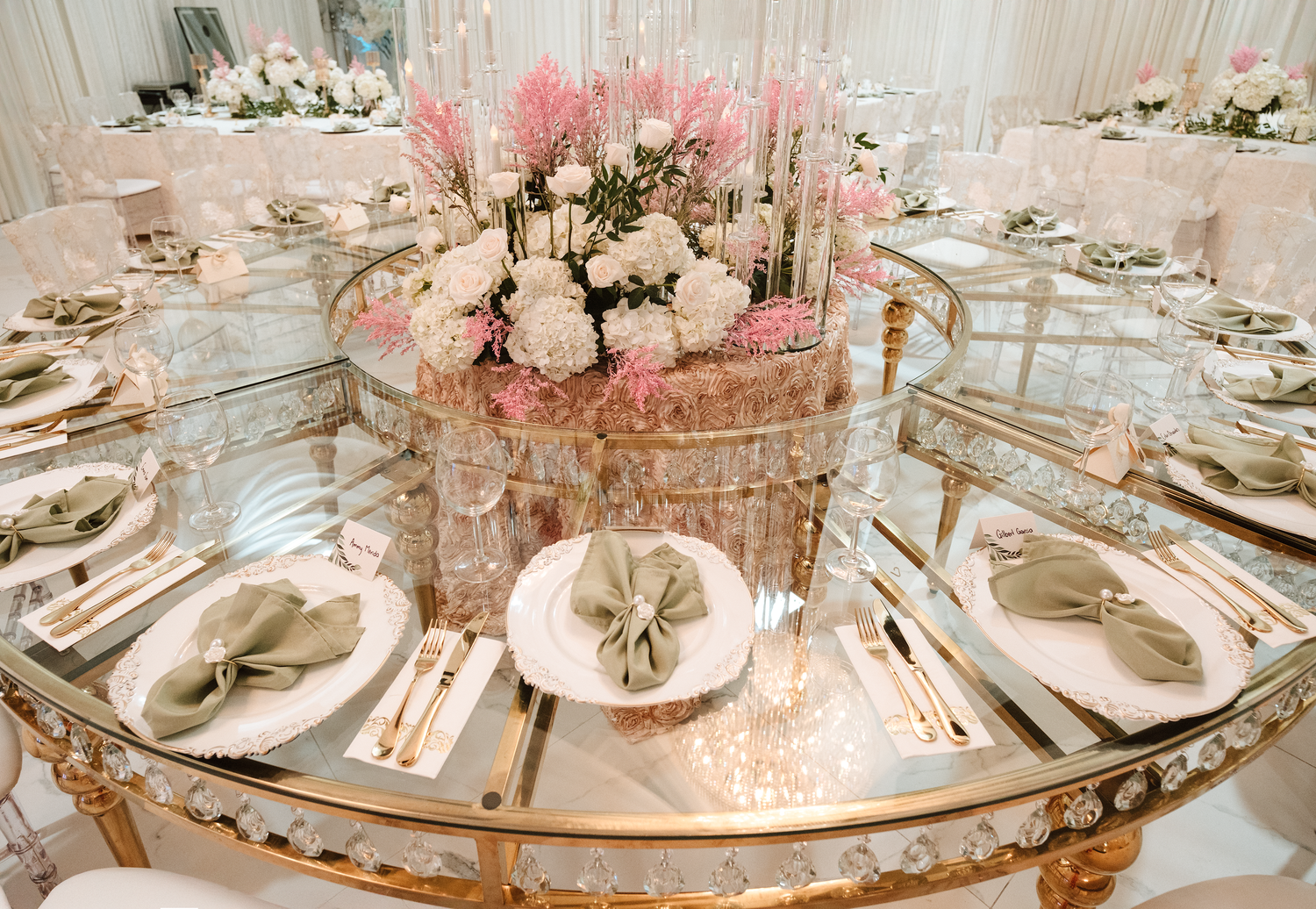 Elegant wedding reception table with gold mirror top, sage green napkins, gold cutlery, and pink floral centerpiece.