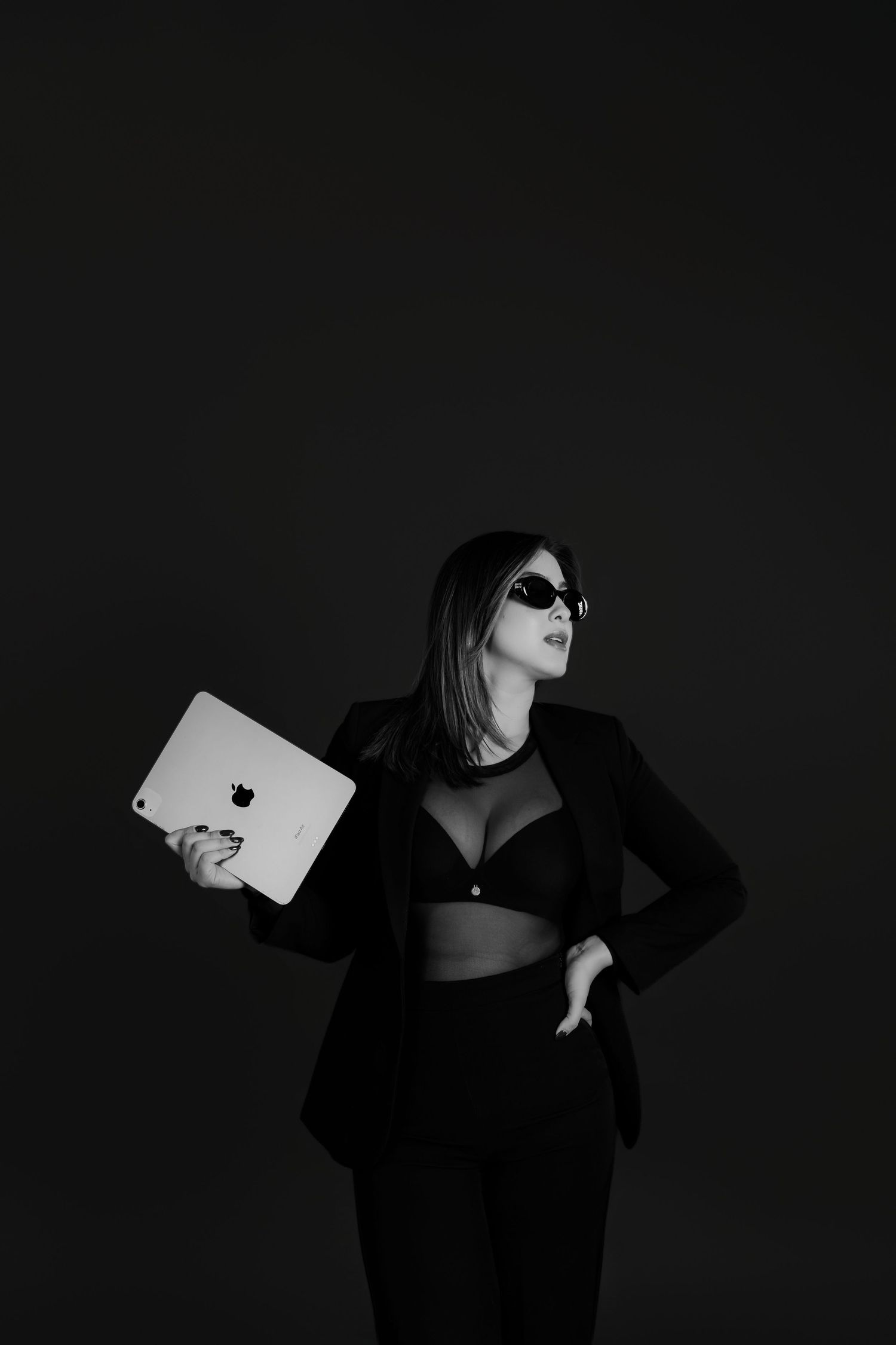 Black and white photo of a stylish woman in sunglasses holding an Apple iPad, wearing a black blazer and bra top.
