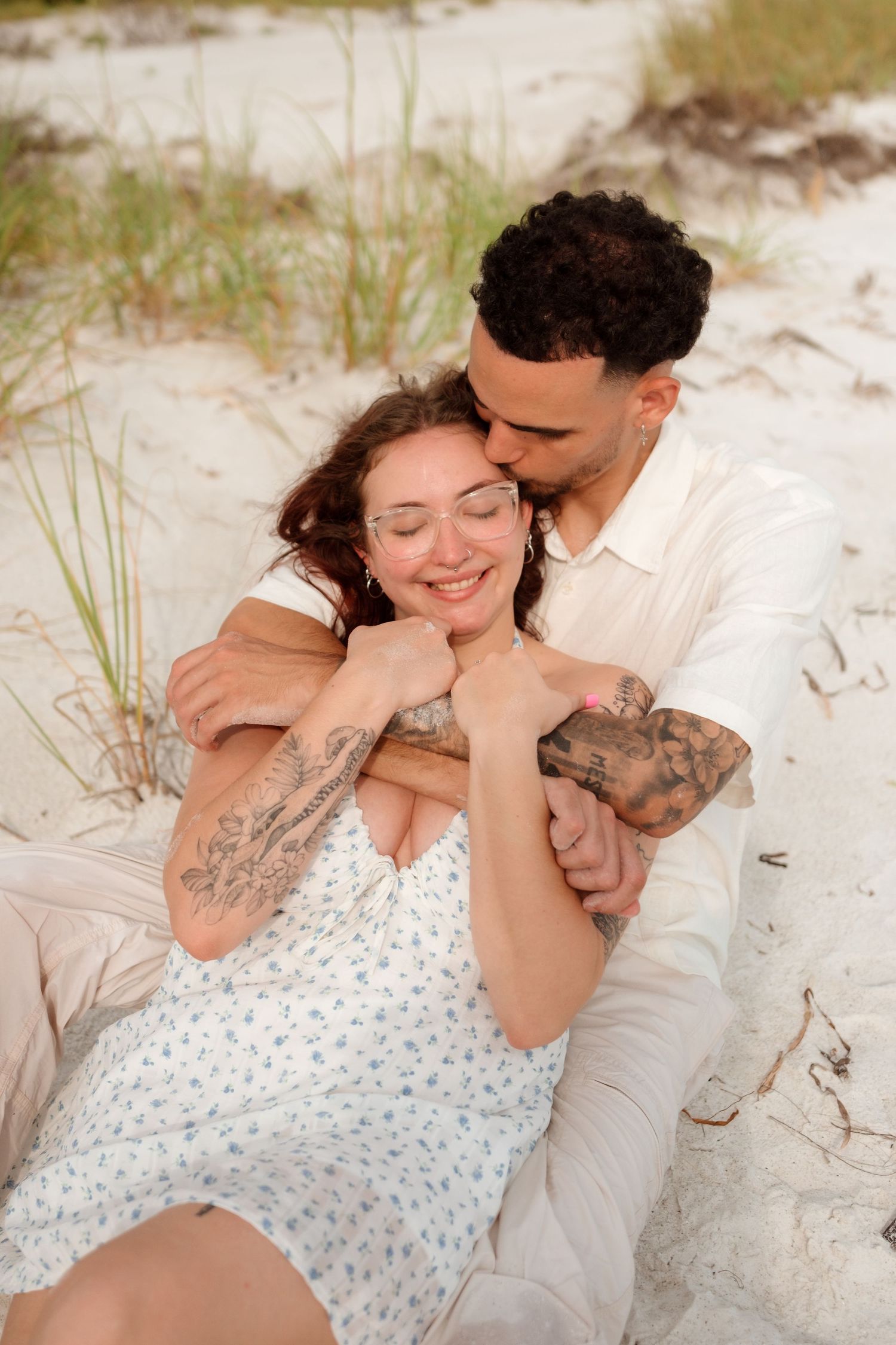 A couple embraces on a sandy beach, the man kissing the woman's head as she smiles, both with tattoos.