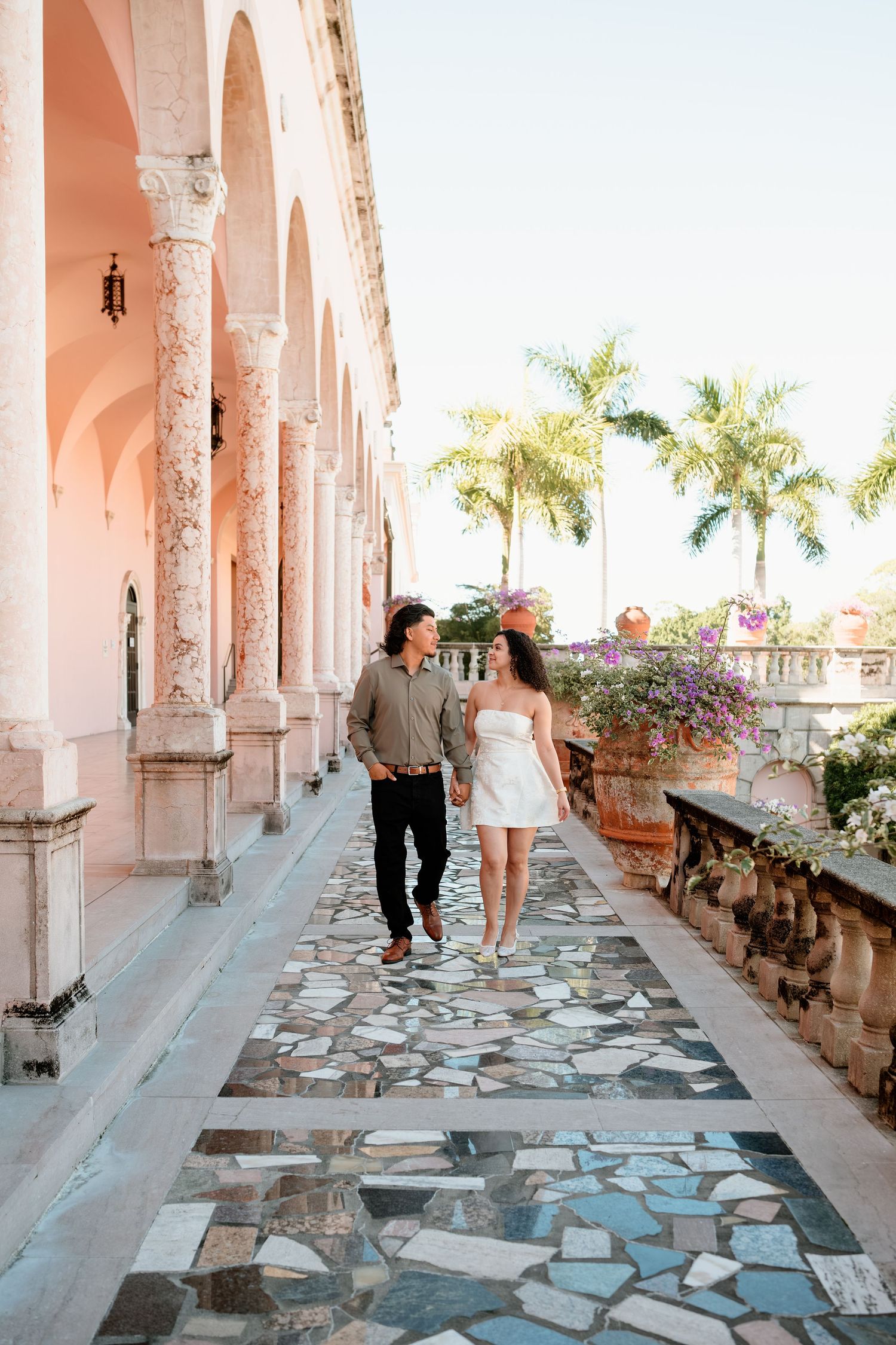 Couple walking hand in hand along elegant pink archway colonnade with palm trees and mosaic stone floor.