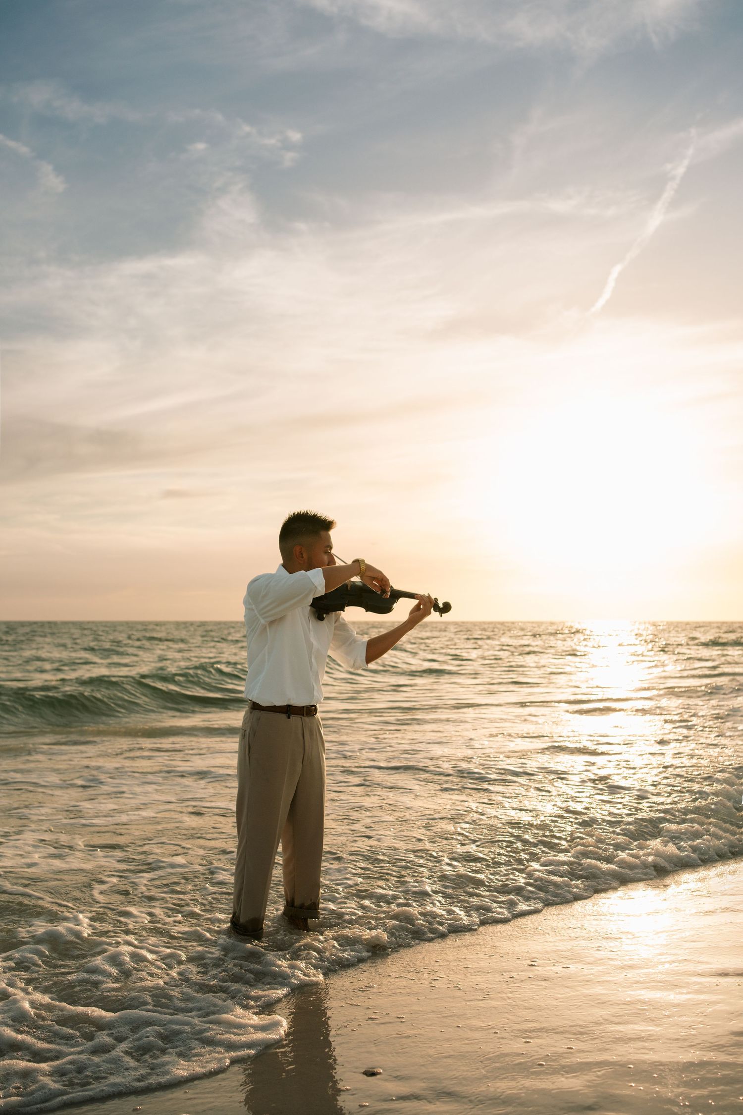 Man playing violin on a beach at sunset, standing in shallow ocean waves wearing white shirt and tan pants.