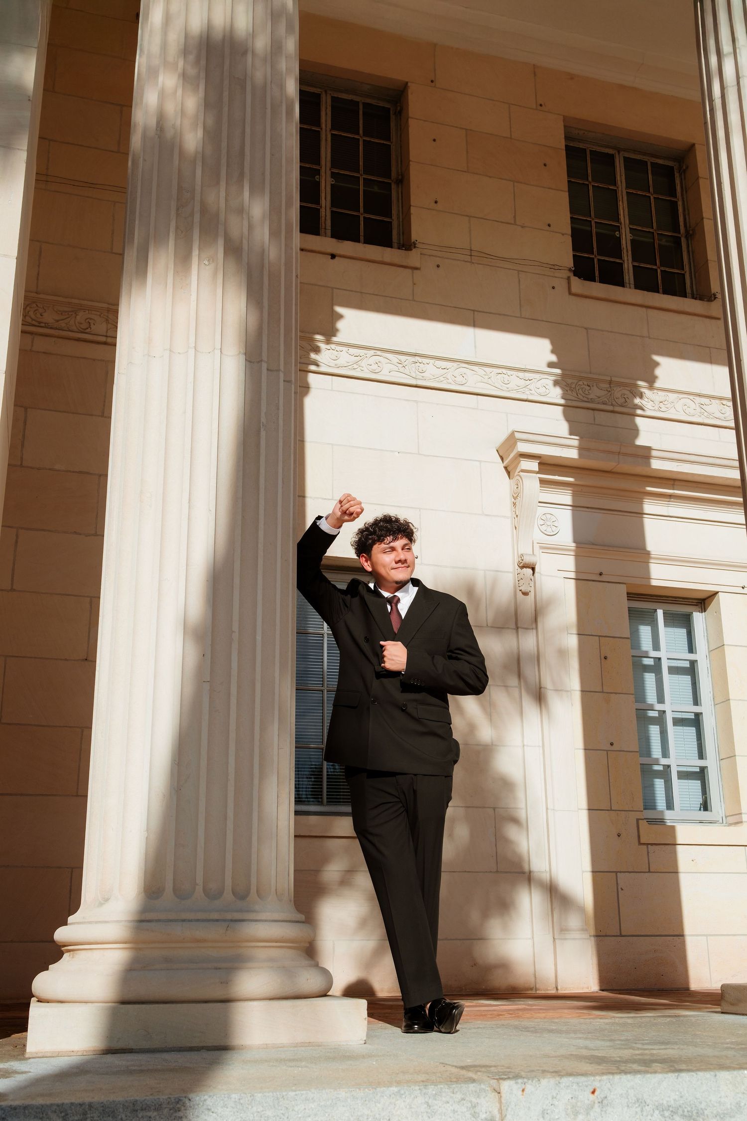 Young man in black suit poses confidently beside large white columns of a classical building in sunlight.