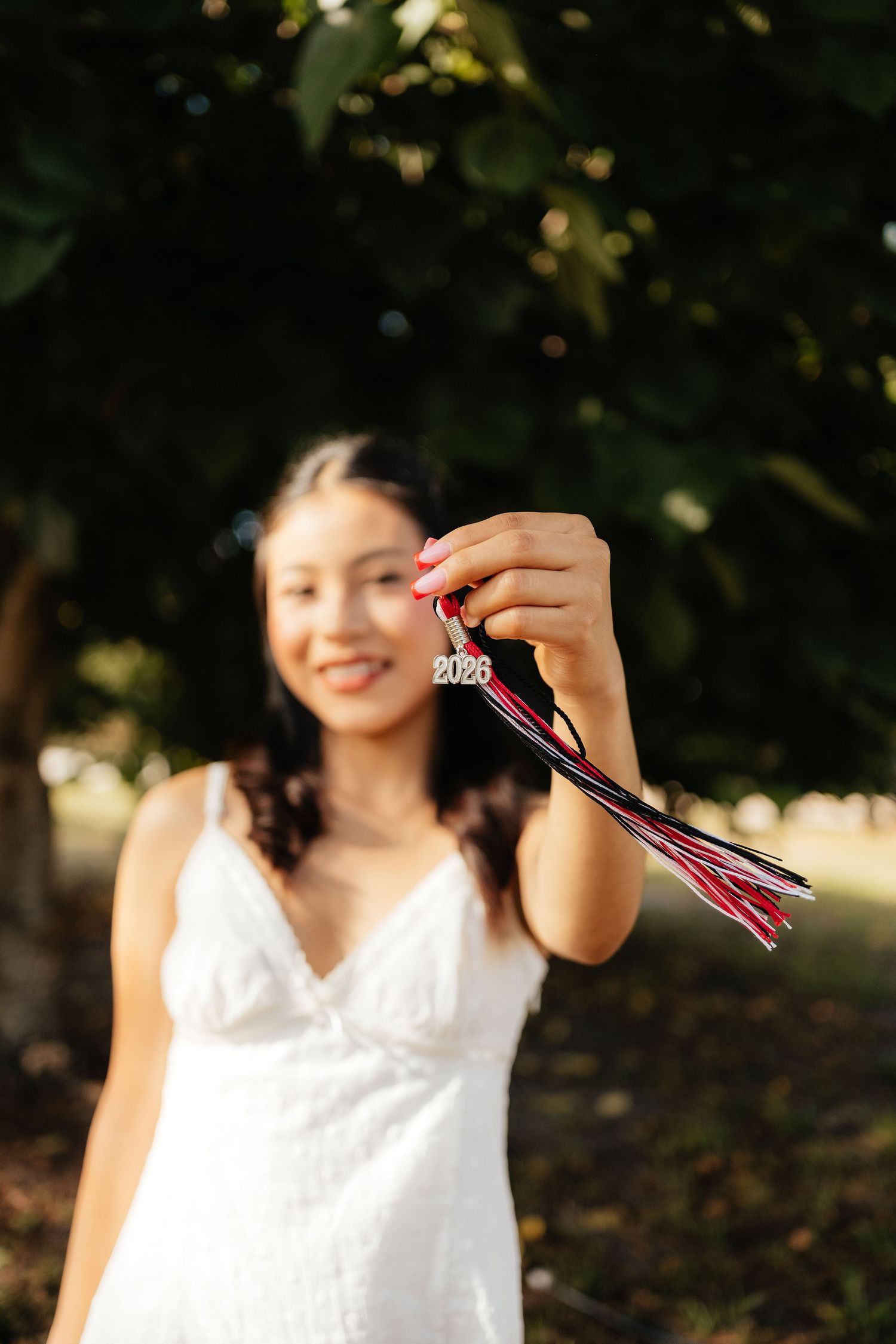 Smiling graduate in white dress holds 2023 tassel toward camera outdoors.
