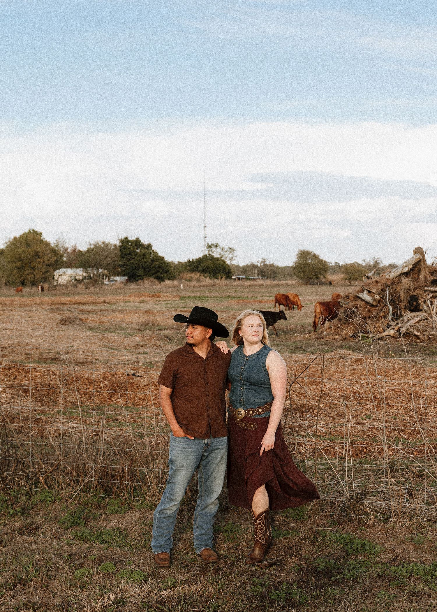 Couple in western attire posing in a rural field with cattle and open sky backdrop.