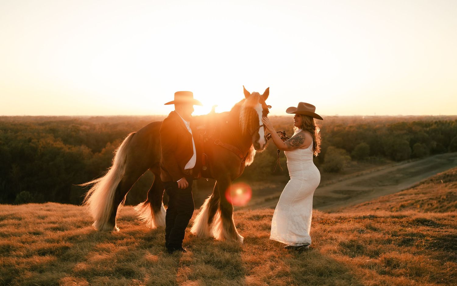 Couple in cowboy hats with a horse at golden sunset on a grassy hill, perfect for rustic wedding inspiration.