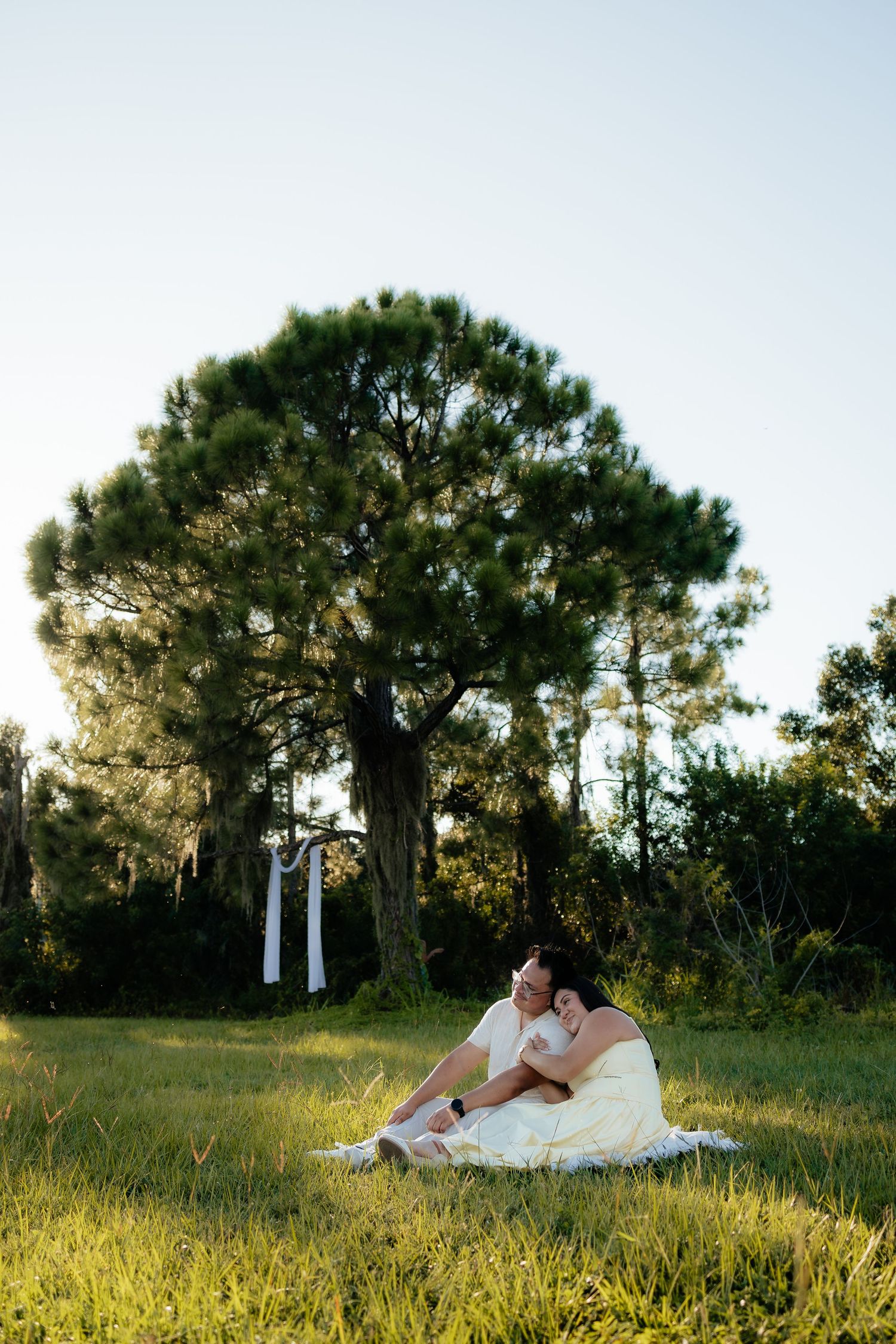 Bride and groom sitting together on grass in front of a large pine tree during golden hour outdoor wedding.